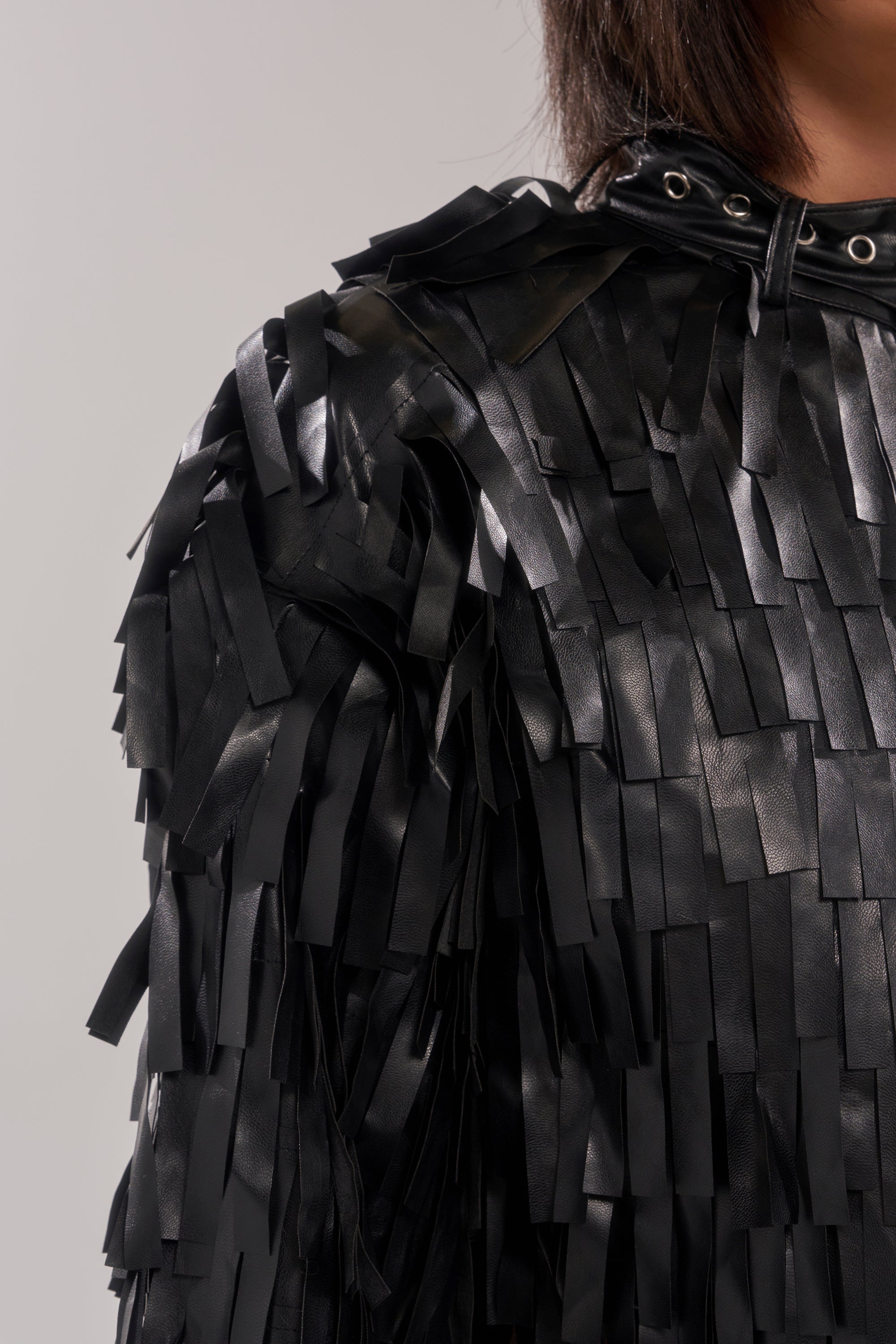 Close-up of a shoulder wearing the RIDE EM FRINGE MOTO—a black faux leather jacket with layered rectangular fringes and metal eyelets at the collar. Its shiny texture pops against a plain light gray background.
