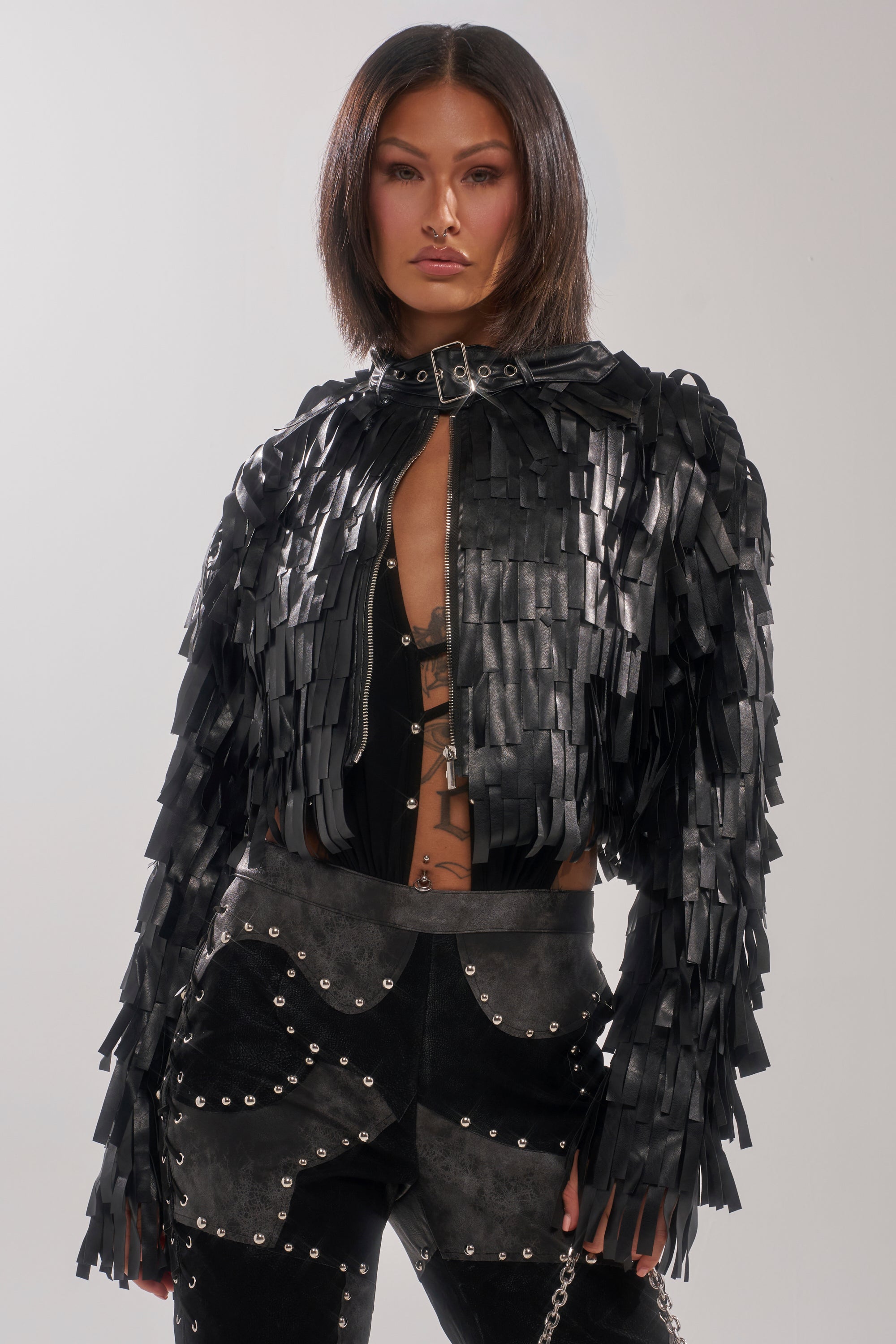 A model poses against a light gray background wearing the RIDE EM FRINGE MOTO—a black moto jacket with fringe and metallic details—styled with a black top and black pants featuring silver studs and chain accents.