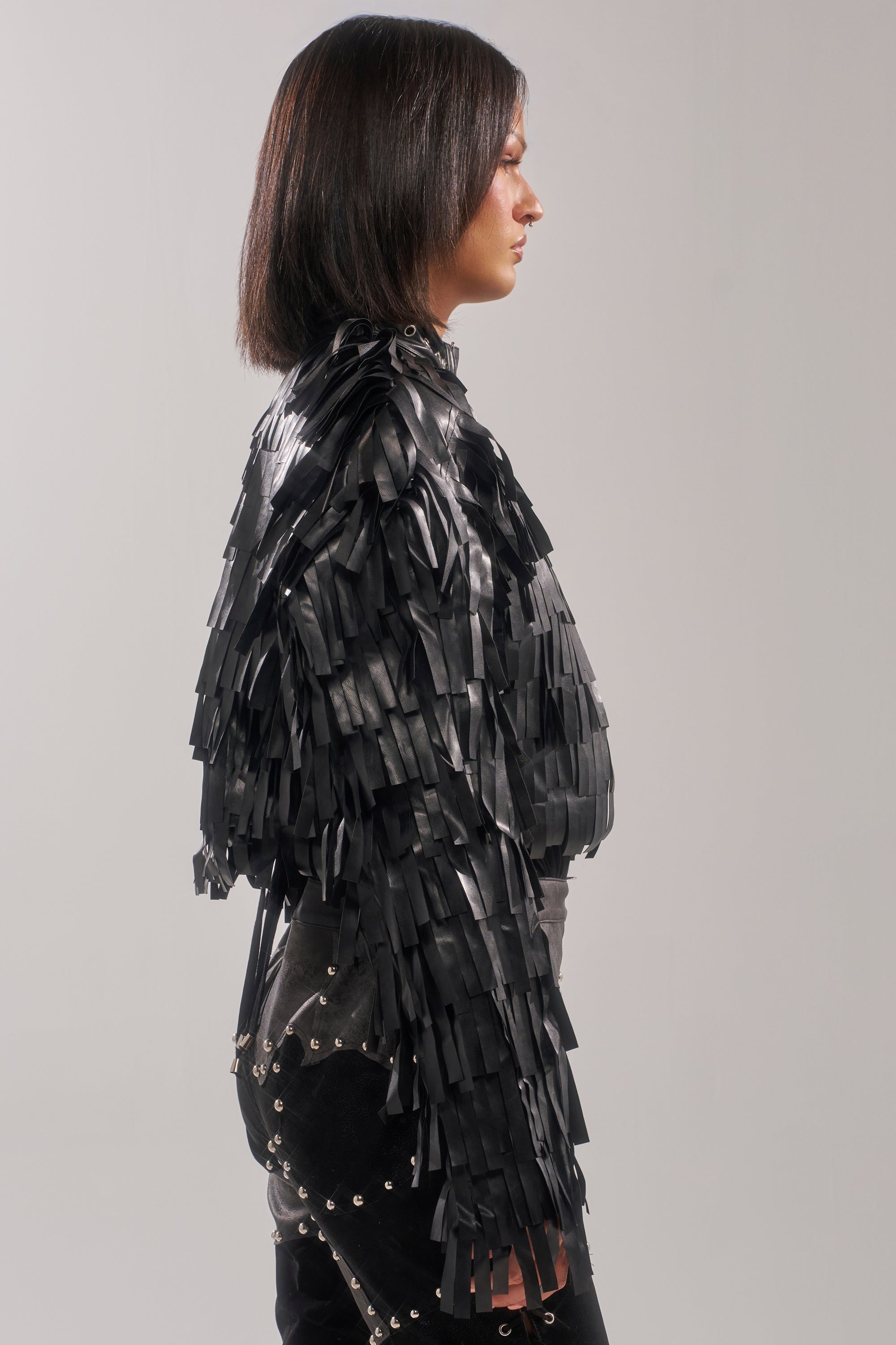 A person with straight, shoulder-length dark hair stands in profile, wearing the RIDE EM FRINGE MOTO jacket and studded black pants against a plain light gray background.