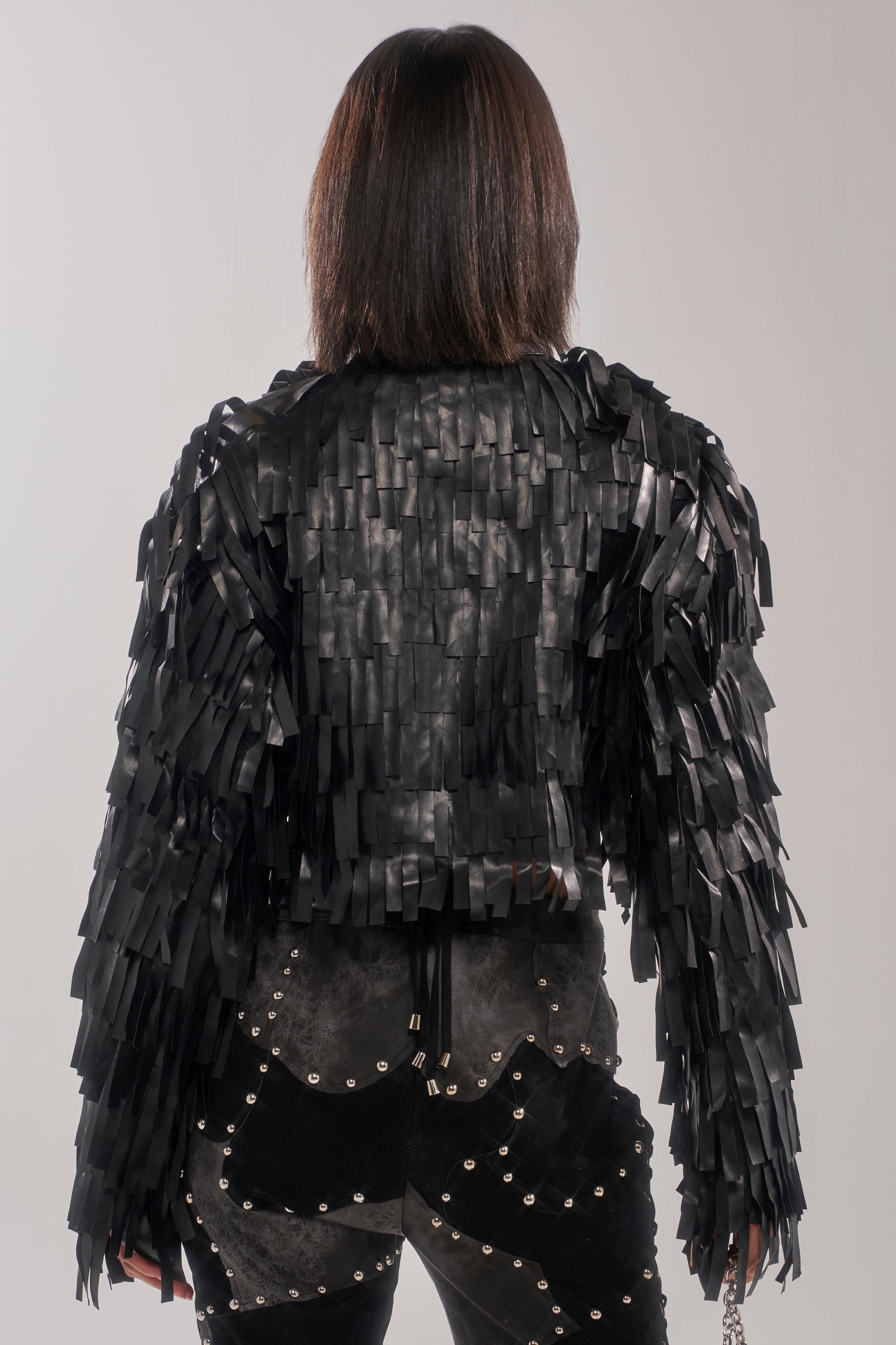 A person with shoulder-length straight dark hair is seen from behind, wearing the RIDE EM FRINGE MOTO jacket featuring textured strips, paired with black pants embellished with metallic studs against a plain light background.