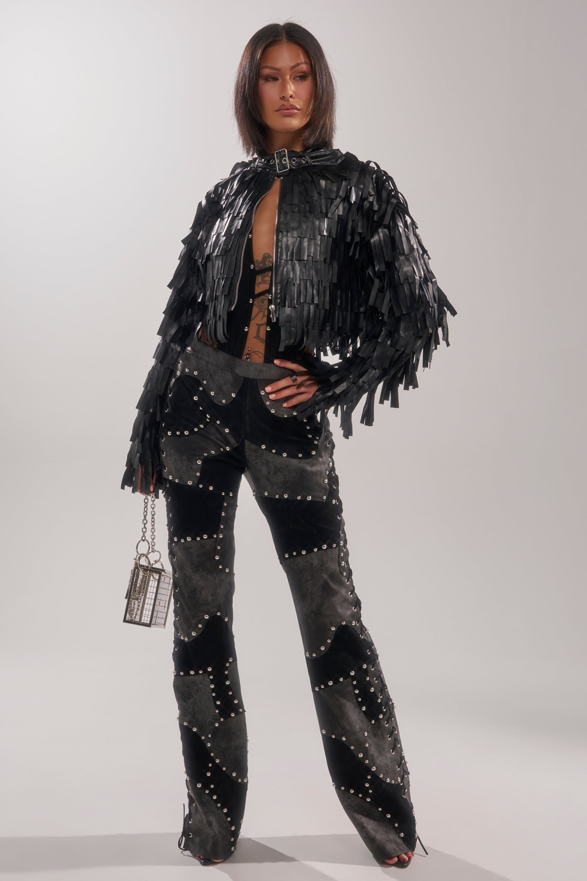 A woman stands confidently in the RIDE EM FRINGE MOTO black jacket, studded black and gray pants, black heels, and holds a small metallic cage purse against a light background.
