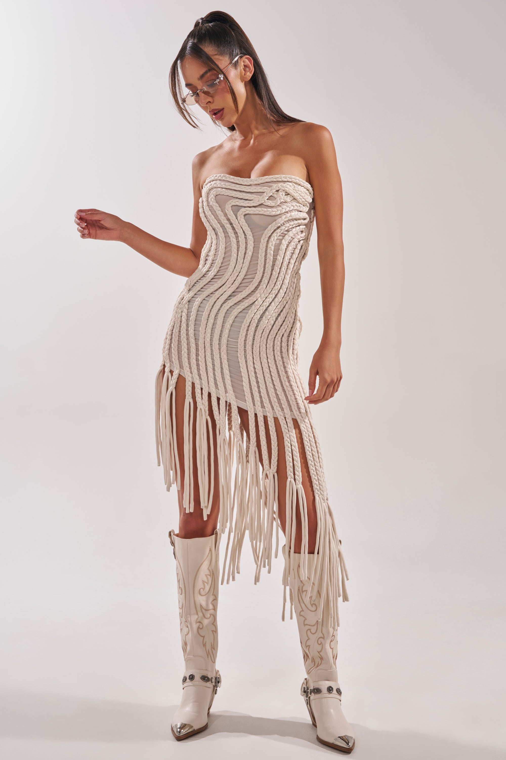 A woman poses in the ALL OVER THE PLACE BRAIDED MIDI DRESS IN IVORY, featuring fringe details and sculptural patterns, paired with knee-high embroidered cowboy boots and silver sunglasses against a plain background. 0