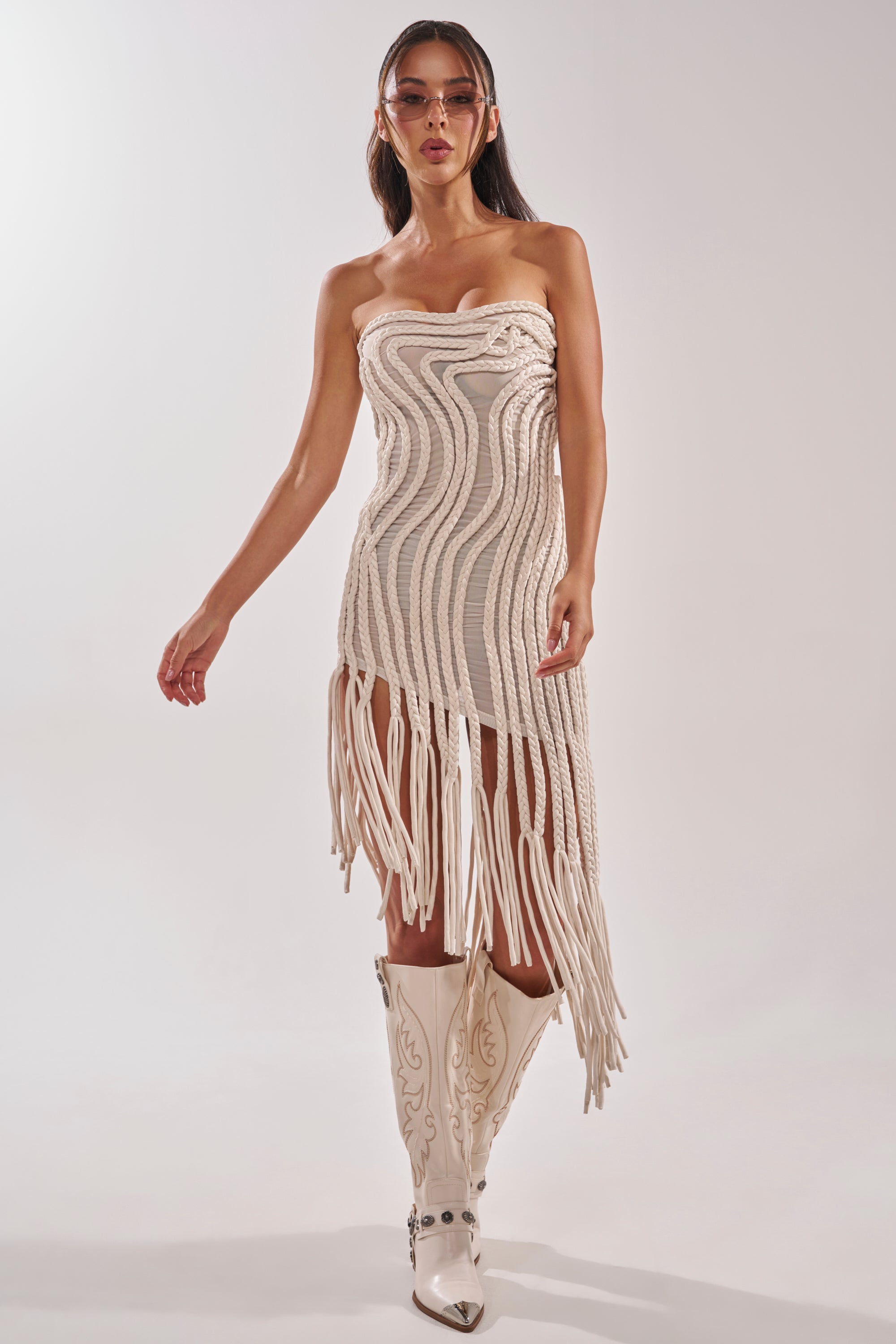 A woman with long brown hair and sunglasses wears the ALL OVER THE PLACE BRAIDED MIDI DRESS IN IVORY, featuring a sculptural wave pattern and long fringe, paired with white cowboy boots against a plain white background. 1
