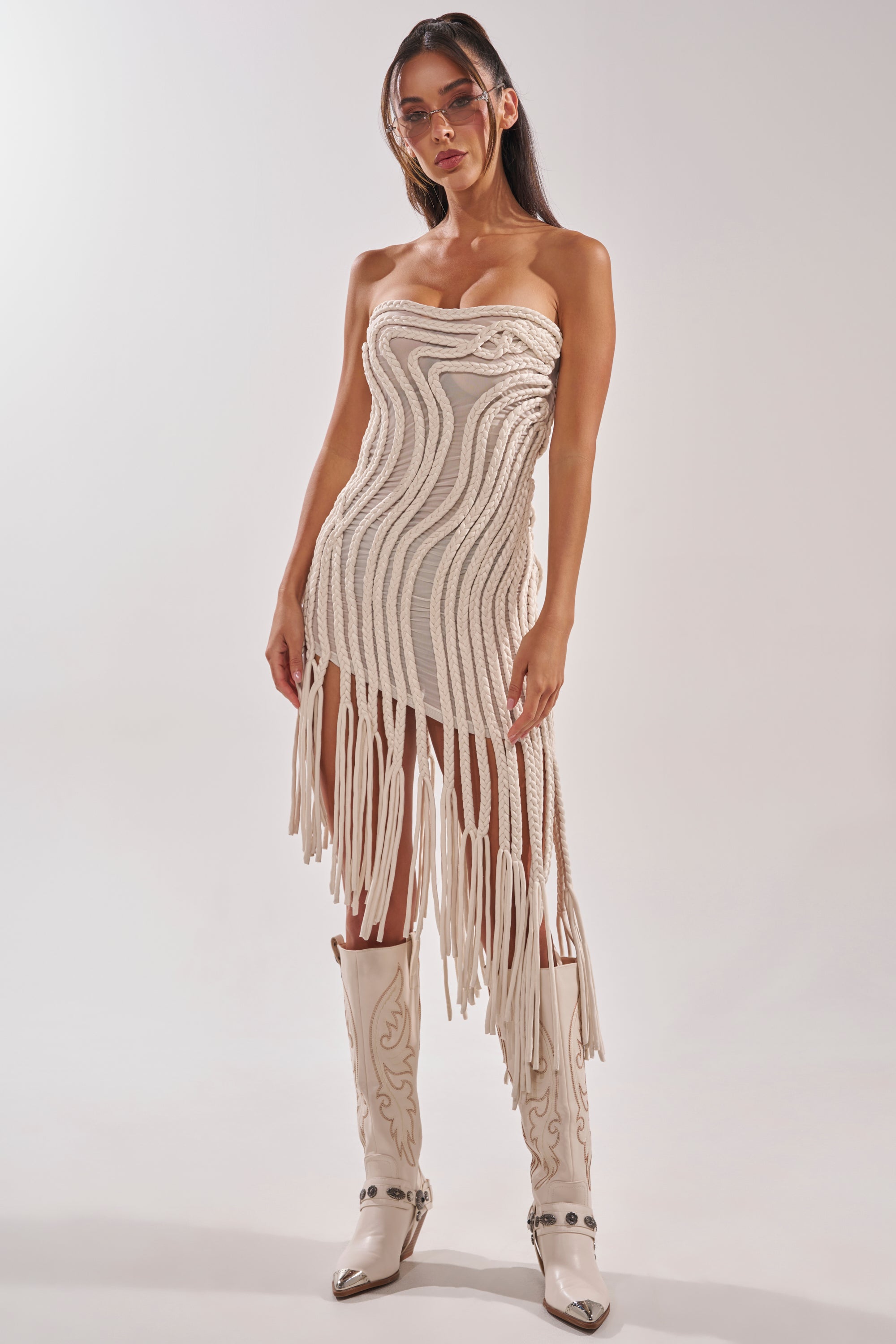 A woman wears the ALL OVER THE PLACE BRAIDED MIDI DRESS IN IVORY, featuring braided details and long fringe, styled with embroidered white cowboy boots and pink sunglasses, posing against a plain light background. 2