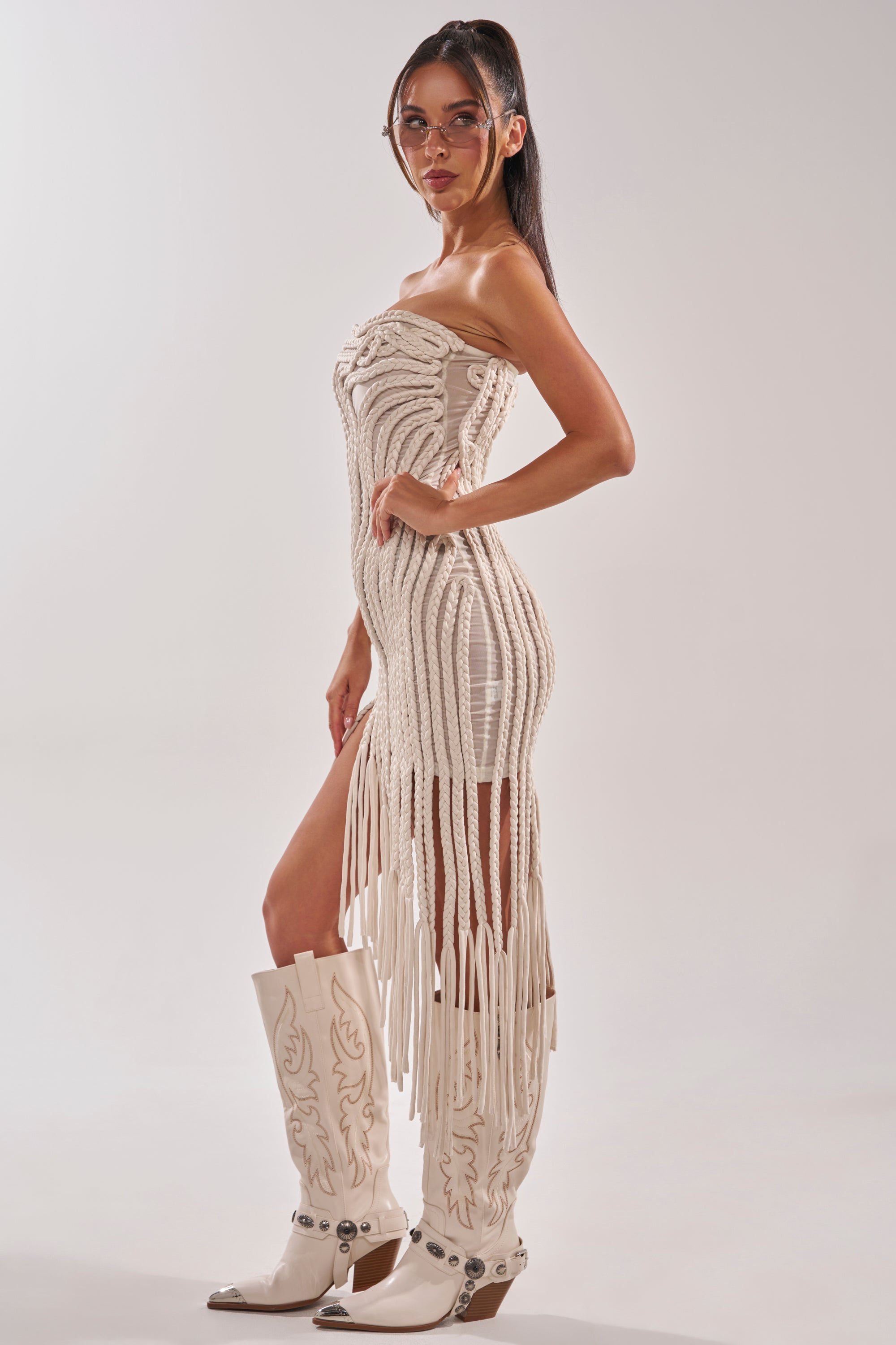 A woman in profile wears the ALL OVER THE PLACE BRAIDED MIDI DRESS IN IVORY and white embroidered cowboy boots. She has long dark hair in a ponytail, glasses, and poses against a plain light background. 4