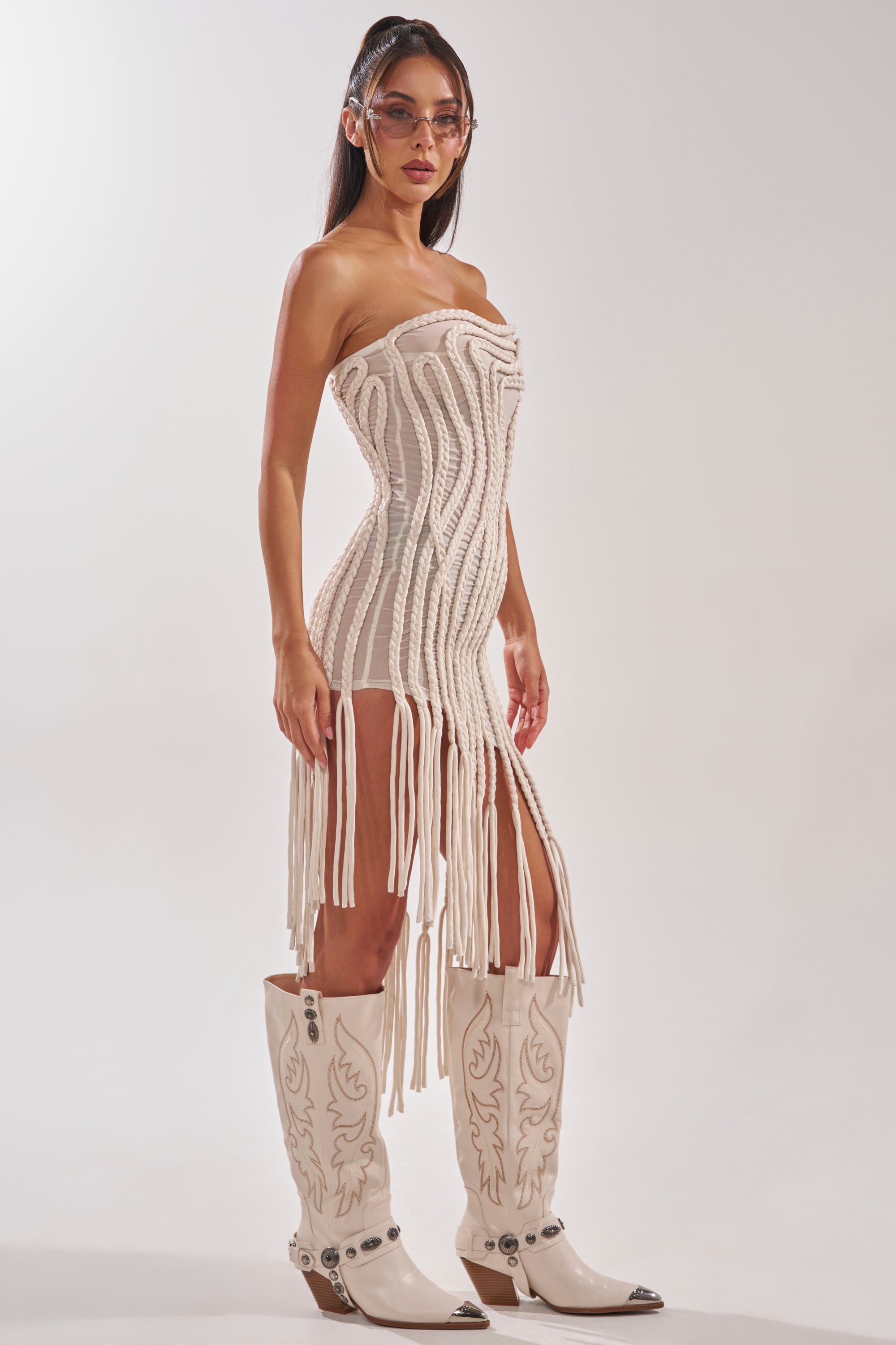A woman in the ALL OVER THE PLACE BRAIDED MIDI DRESS IN IVORY poses against a plain light background, styled with white embroidered cowboy boots, sunglasses, and long hair. 5