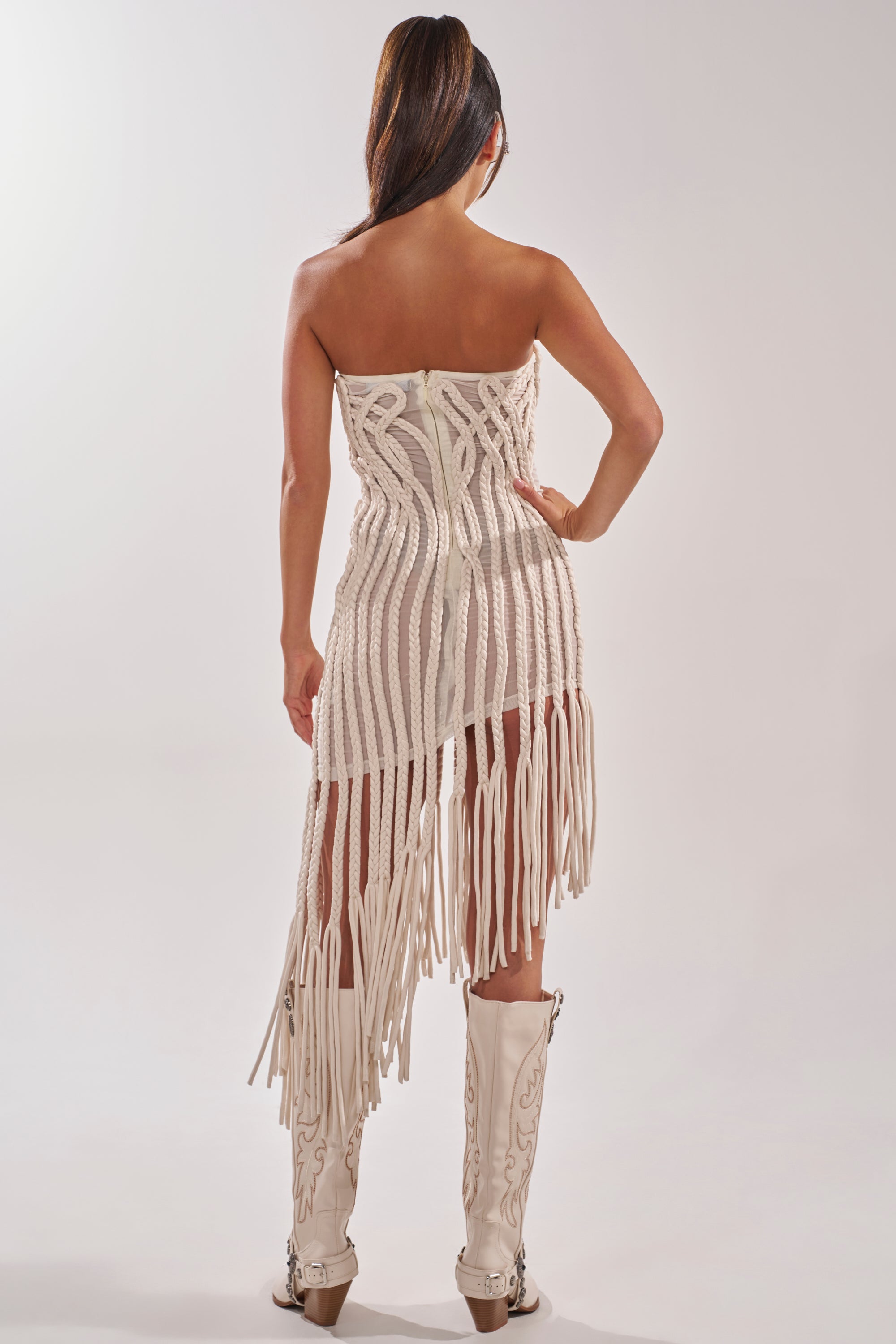 A woman faces away, wearing the ALL OVER THE PLACE BRAIDED MIDI DRESS IN IVORY and knee-high white embroidered cowboy boots. Her long hair is styled in a sleek ponytail. 6