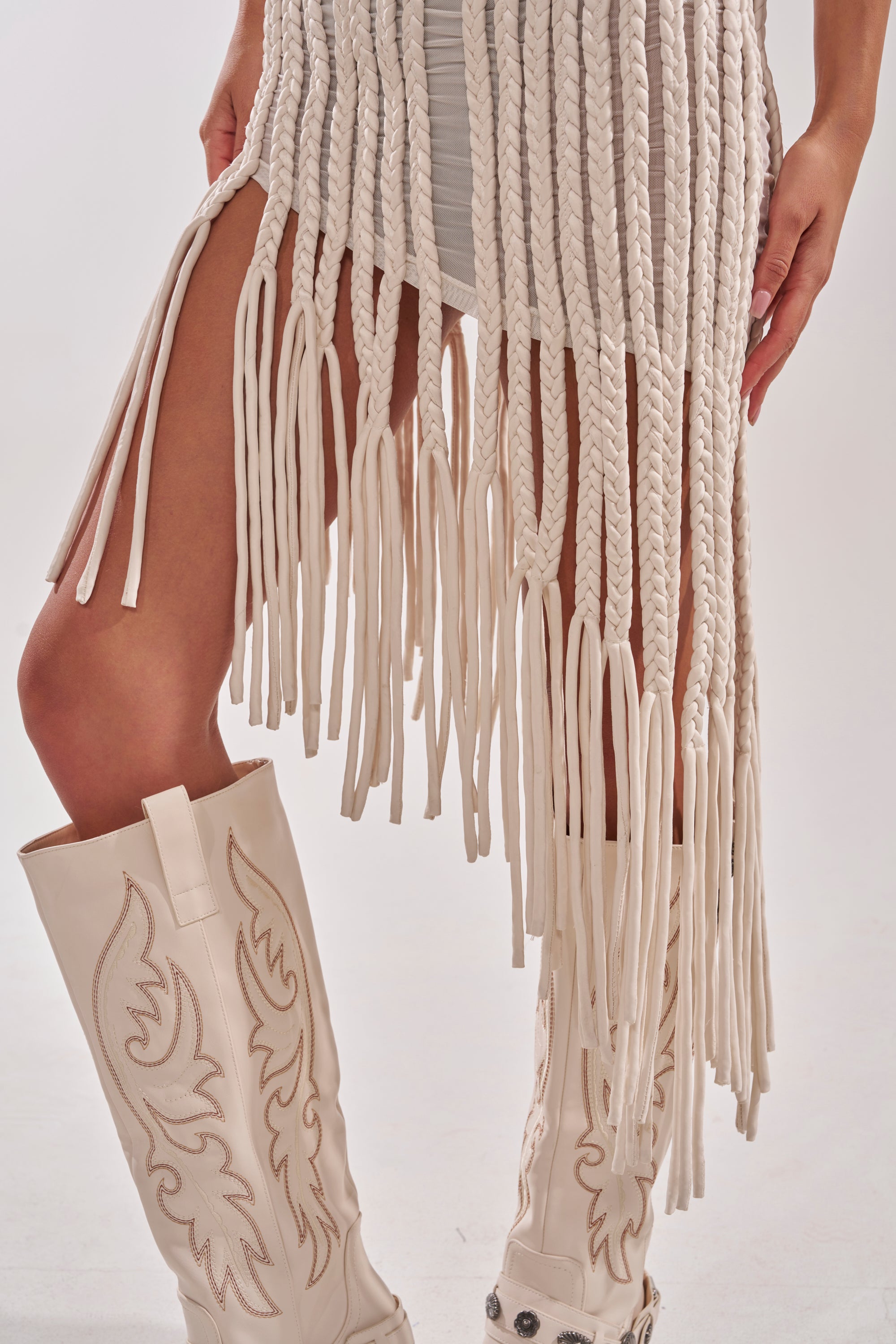 Someone stands against a plain light background wearing the ALL OVER THE PLACE BRAIDED MIDI DRESS IN IVORY, a cream textured midi dress with long fringe tassels, paired with white embroidered cowboy boots. 8