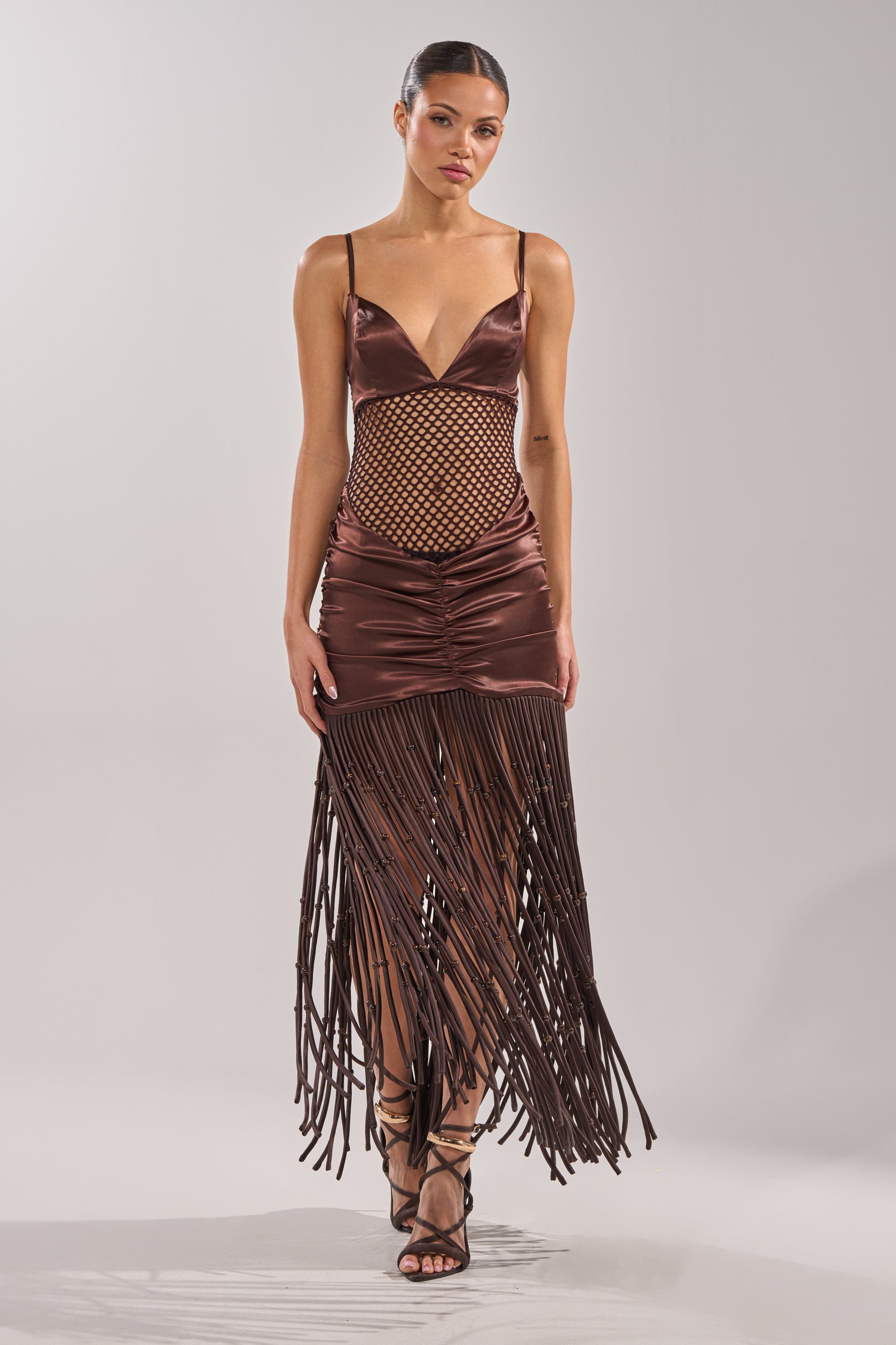 A woman with slicked-back hair wears the MIDNIGHT MOCHA MAXI DRESS, a sleeveless satin maxi with a deep V-neck, fishnet midsection, and long fringe accents, styled with strappy high-heeled sandals. 2