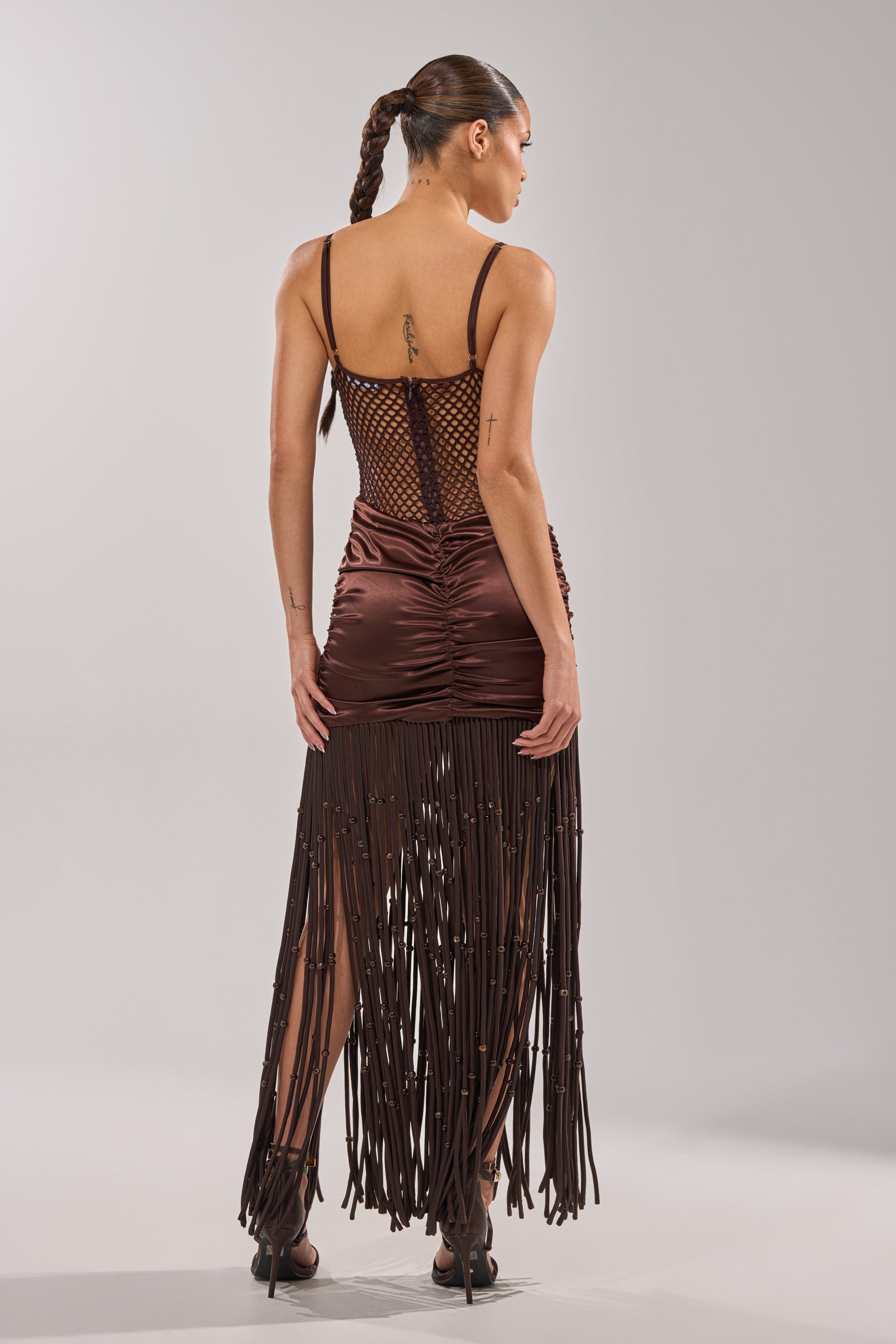 A woman with a long braided ponytail models the MIDNIGHT MOCHA MAXI DRESS, showcasing its netted top, ruched back, and long satin fringe skirt. She stands in a neutral studio with her back to the camera, hands at her sides. 5