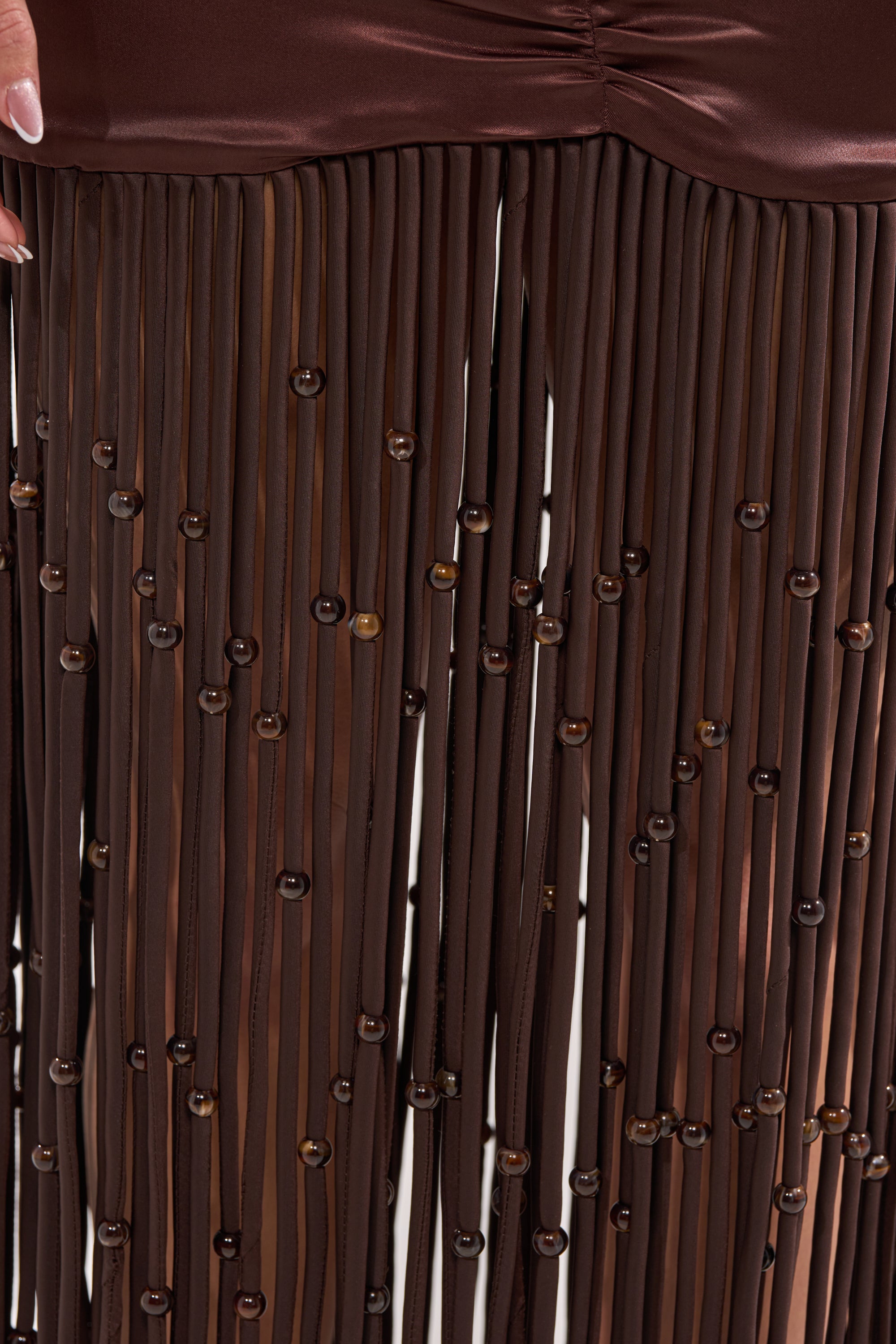 Close-up of long, vertical fringe strips with small, round beads on a brown skirt. A manicured hand appears at the edge—an ideal accent piece to pair with the MIDNIGHT MOCHA MAXI DRESS for evening events. 7