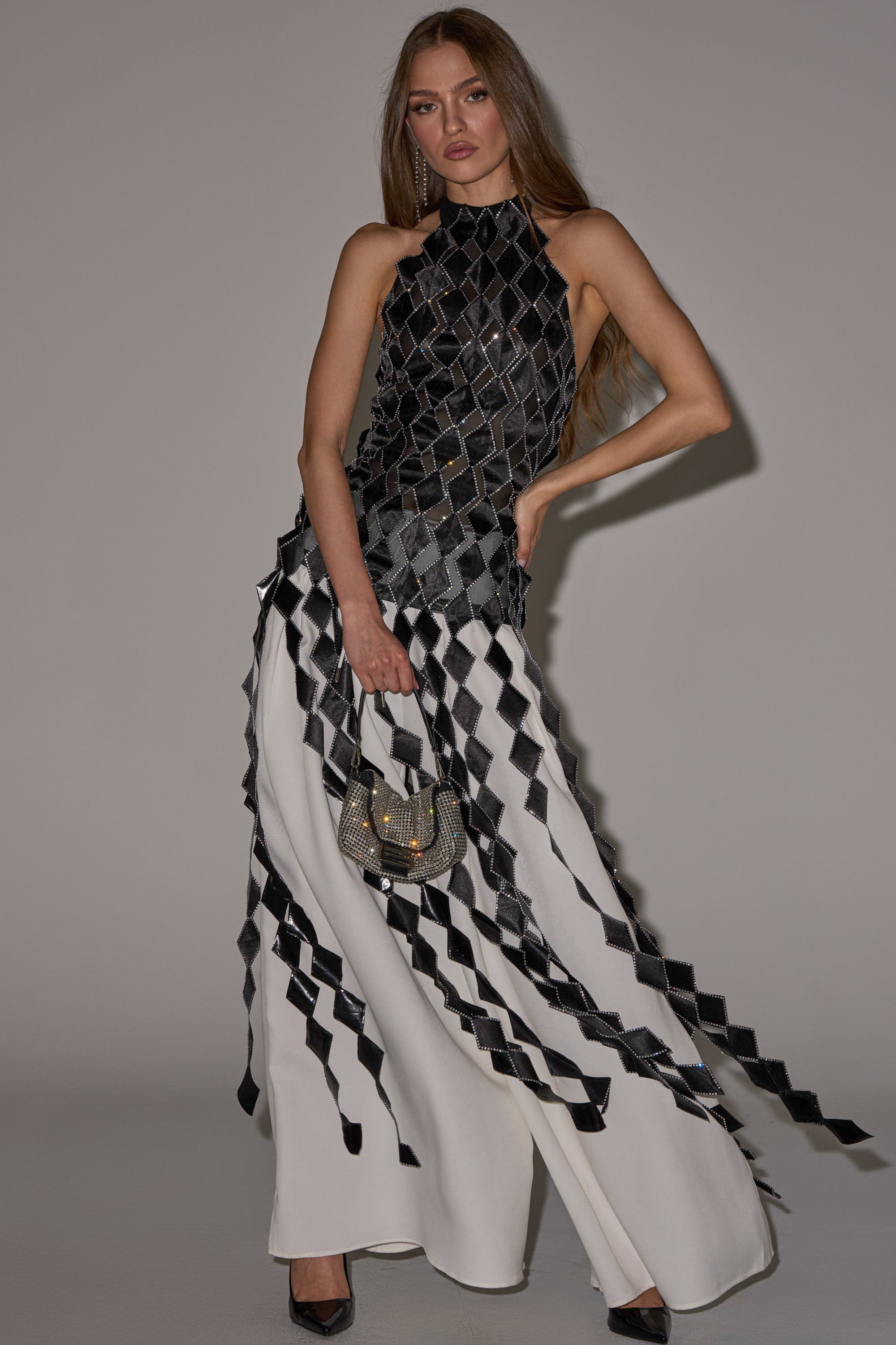 A woman wears the ELINA SATIN TOP under a sleeveless, black and white geometric-patterned dress with a high neckline and layered diamond shapes, styled with a small handbag and black heels. 0