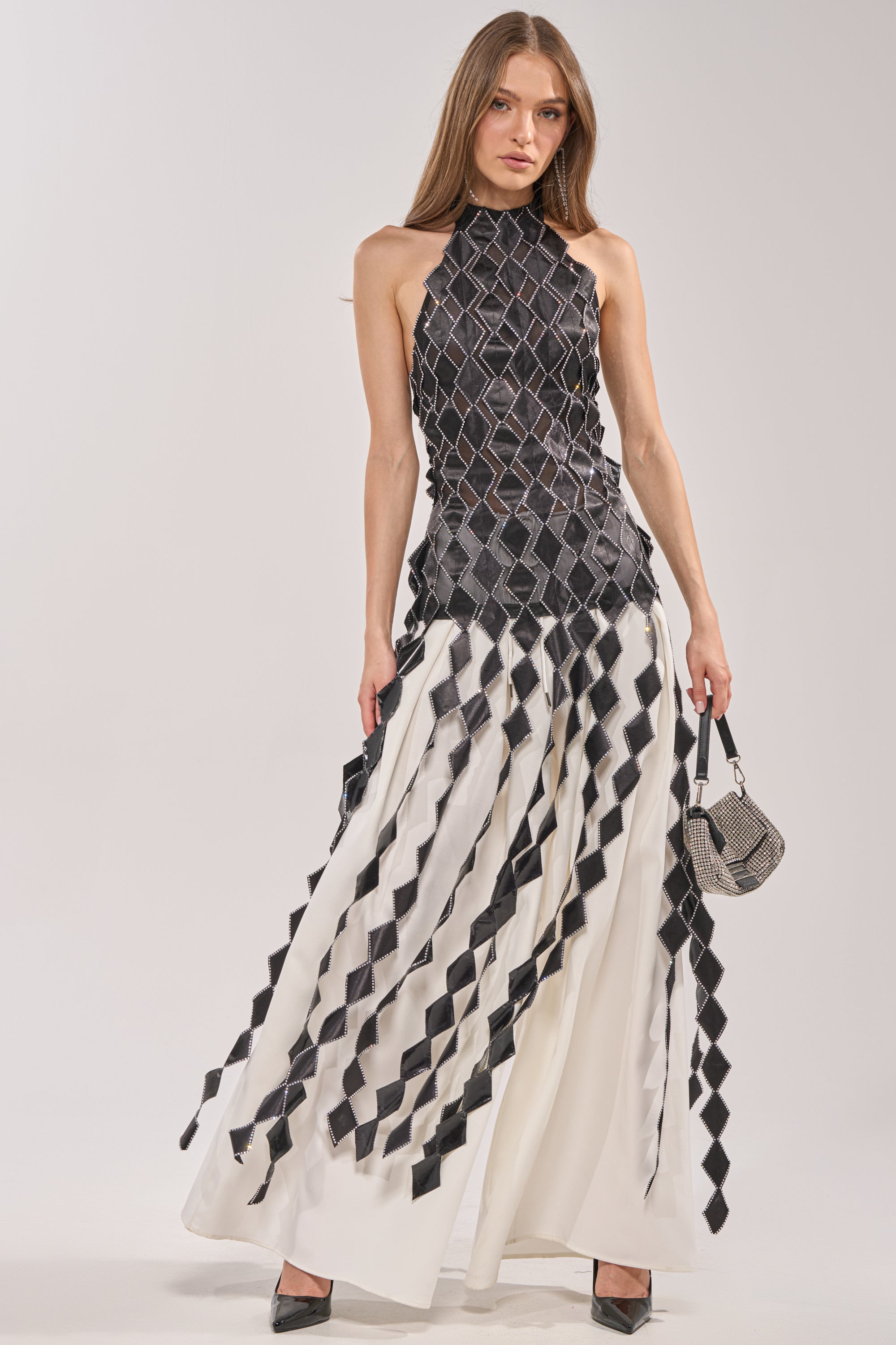 A woman stands holding a small handbag while wearing the ELINA SATIN TOP, a sleeveless black-and-white geometric top featuring diamond-shaped strips and subtle rhinestone trim over a long white skirt. 1