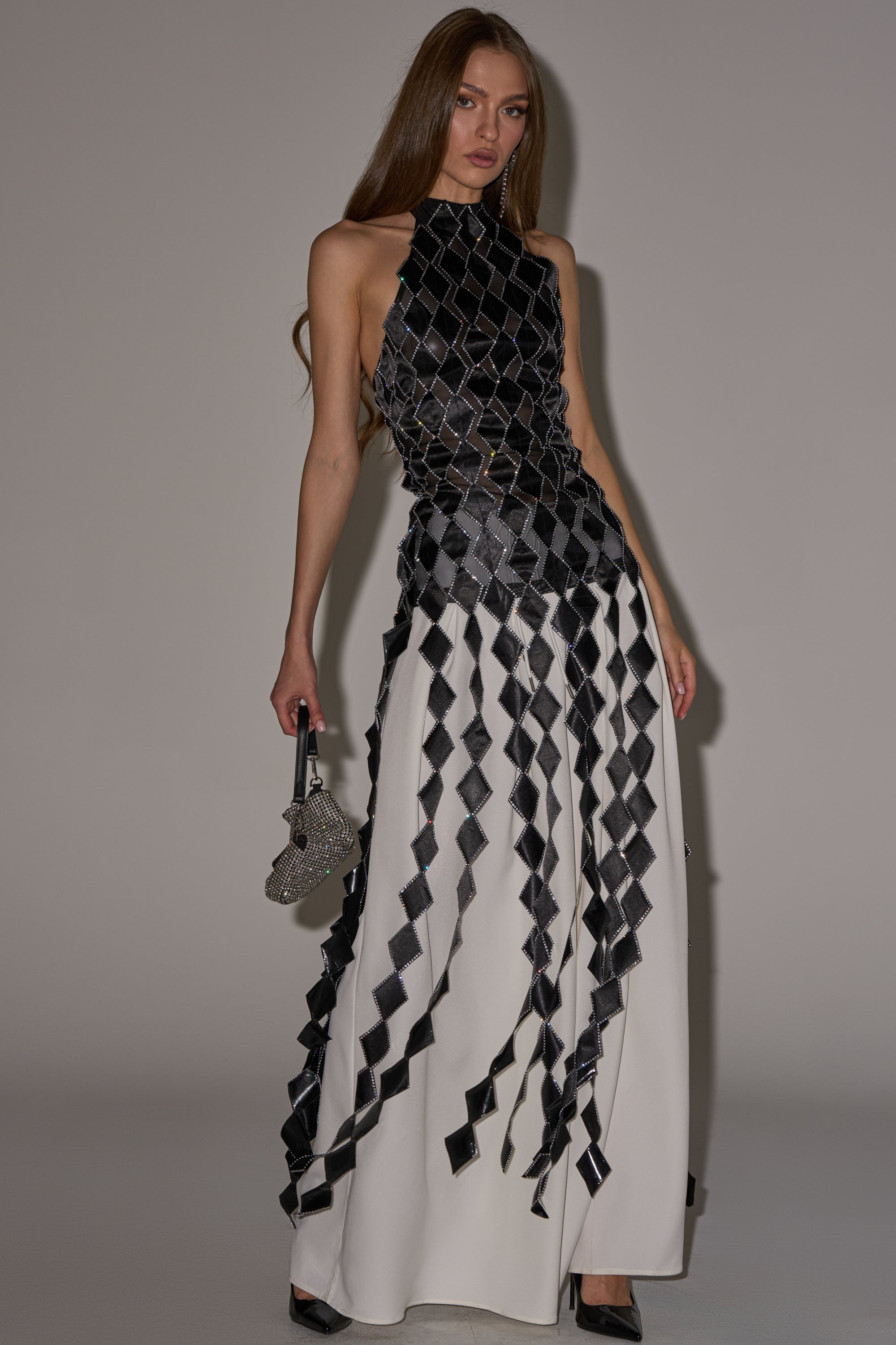 A woman with long hair wears the ELINA SATIN TOP, a sleeveless mesh halter, paired with flowing white pants featuring a black geometric diamond pattern. She holds a small handbag and poses against a plain background. 2