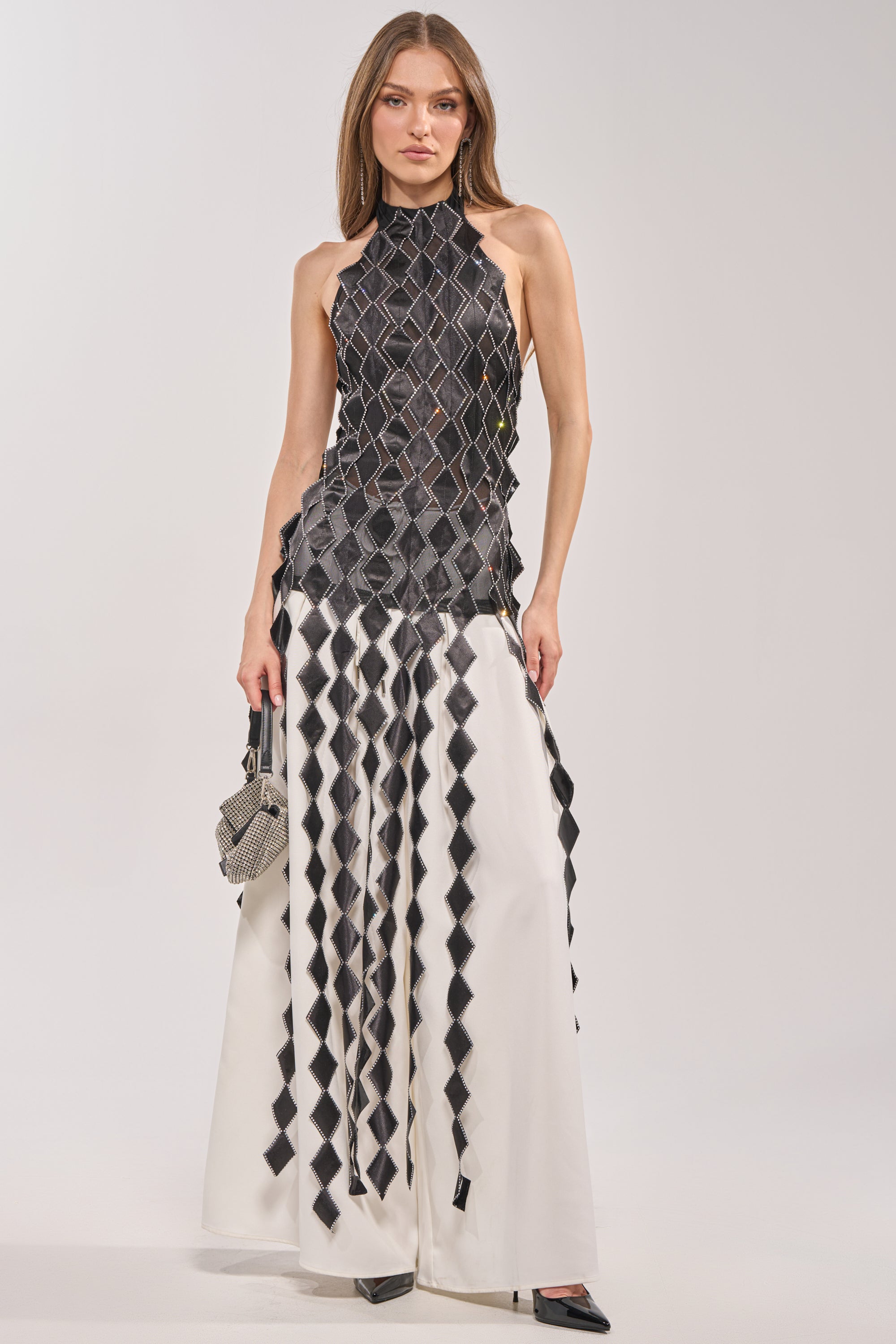 Wearing the ELINA SATIN TOP, a woman pairs the longline black diamond-patterned top with white wide-leg pants featuring matching diamonds. She accessorizes with a small handbag, black heels, and subtle rhinestone trim for an elegant finish. 3
