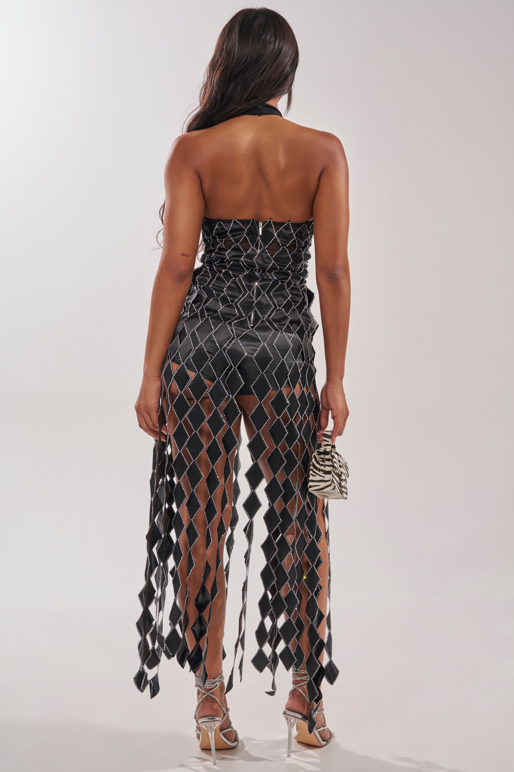 A woman with long dark hair, facing away from the camera, wears the ELINA SATIN TOP paired with a strapless black dress featuring diamond-shaped cutouts and rhinestone trim. She holds a zebra-print bag and wears silver high heels. 4