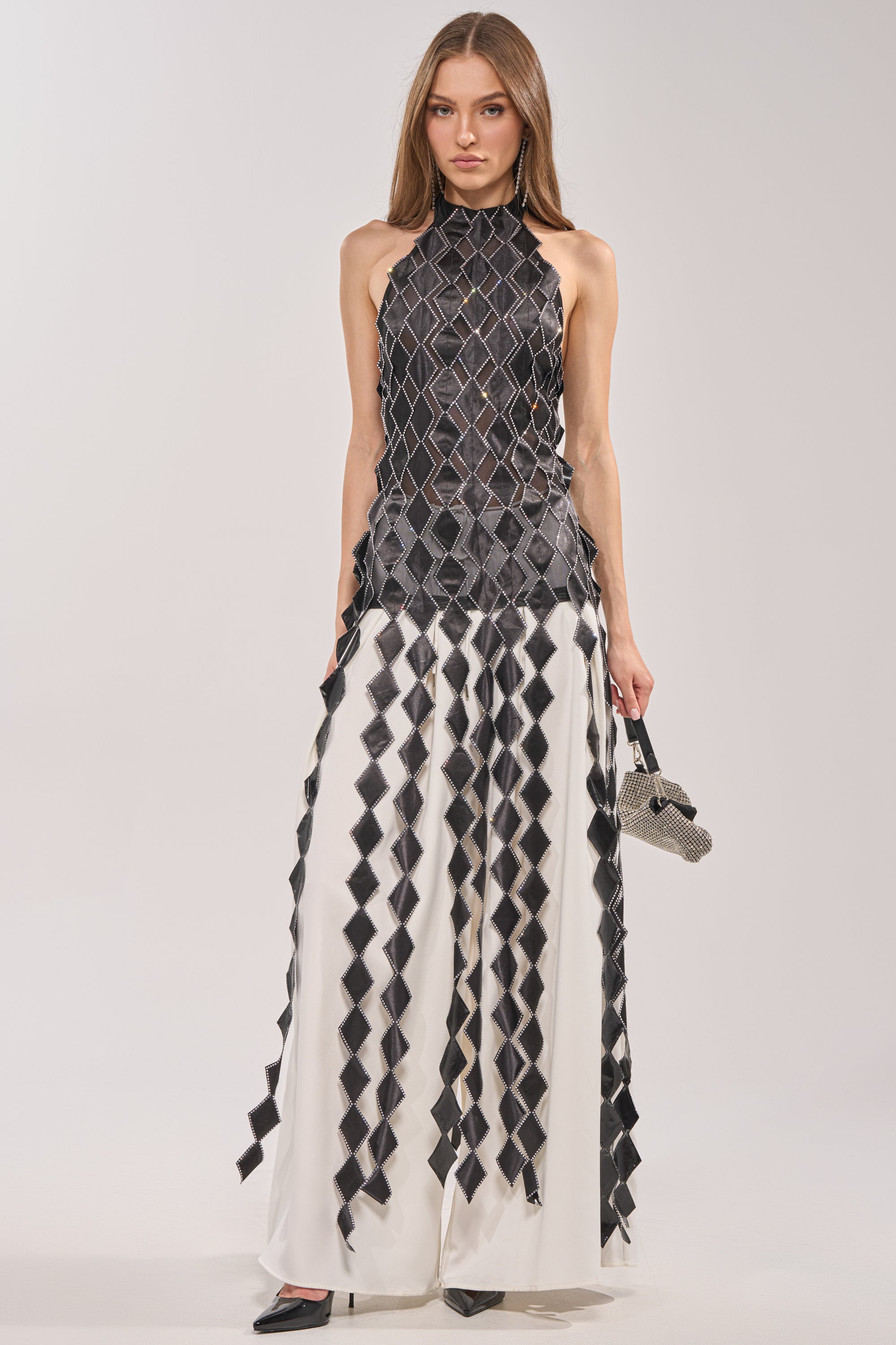 A woman wears the ELINA SATIN TOP, a sleeveless black and white geometric halter with rhinestone trim, paired with matching wide-leg pants. She carries a textured handbag and wears black pointed shoes against a plain backdrop. 4