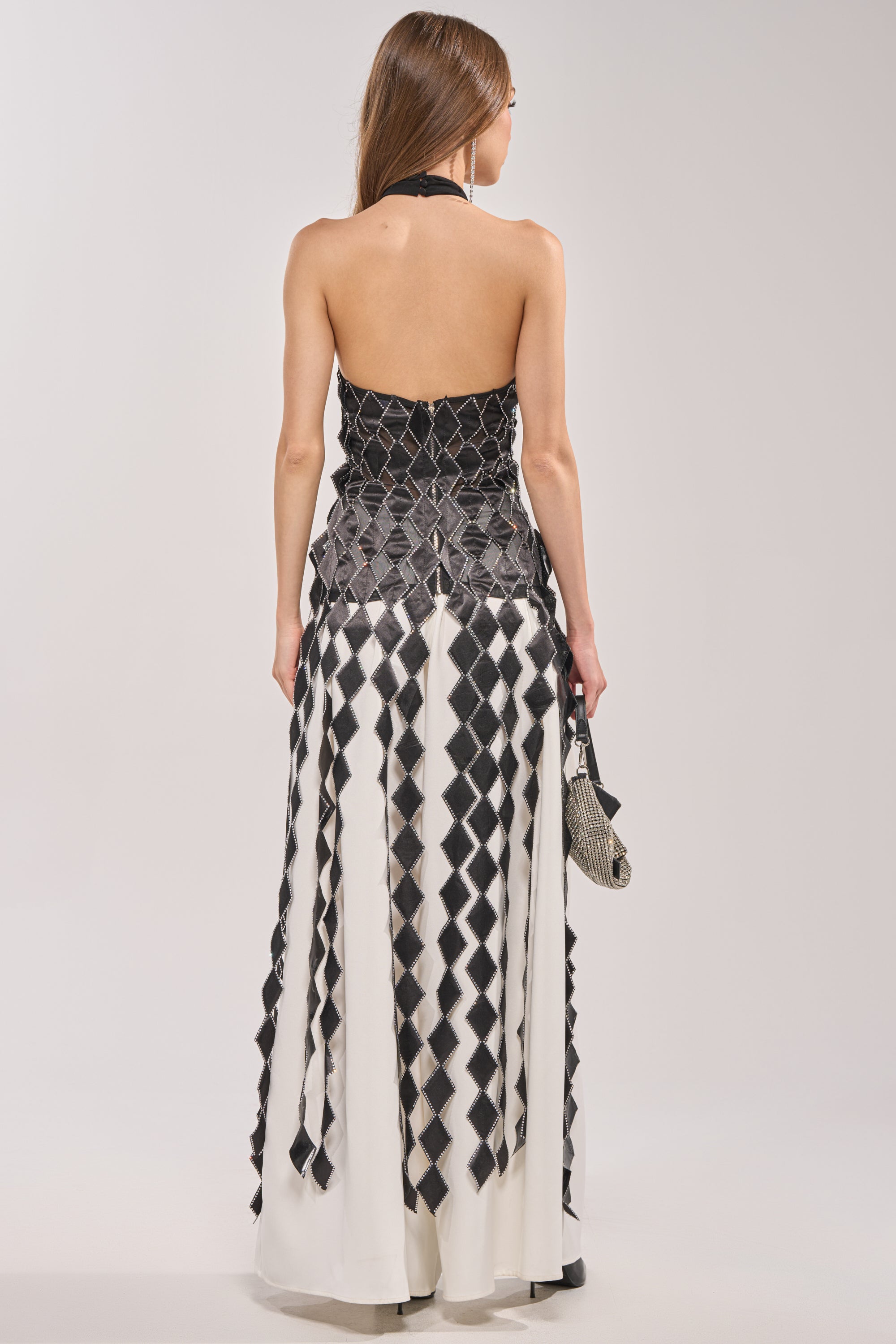 A woman with long hair stands facing away, wearing the ELINA SATIN TOP in a black and white geometric pattern with a strapless design and rhinestone trim. She pairs it with a small black and white clutch in her right hand. 6