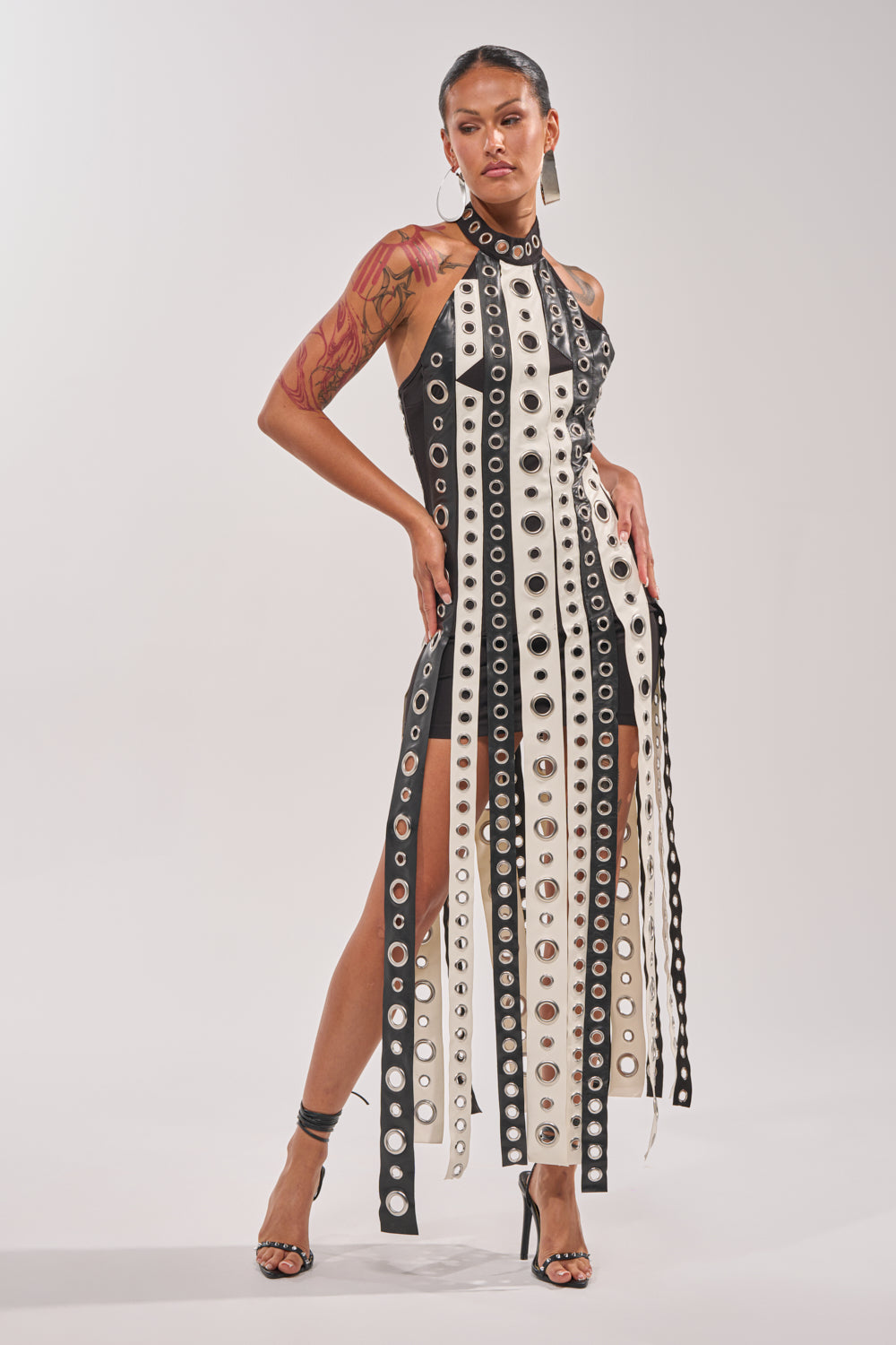 A woman stands confidently in the ON MY BEST BEHAVIOR TASSEL MAXI DRESS, featuring black and white vertical strips with metal eyelets. She’s styled with high heels, hoop earrings, a sleek pulled-back hairstyle, and a visible arm tattoo. 0