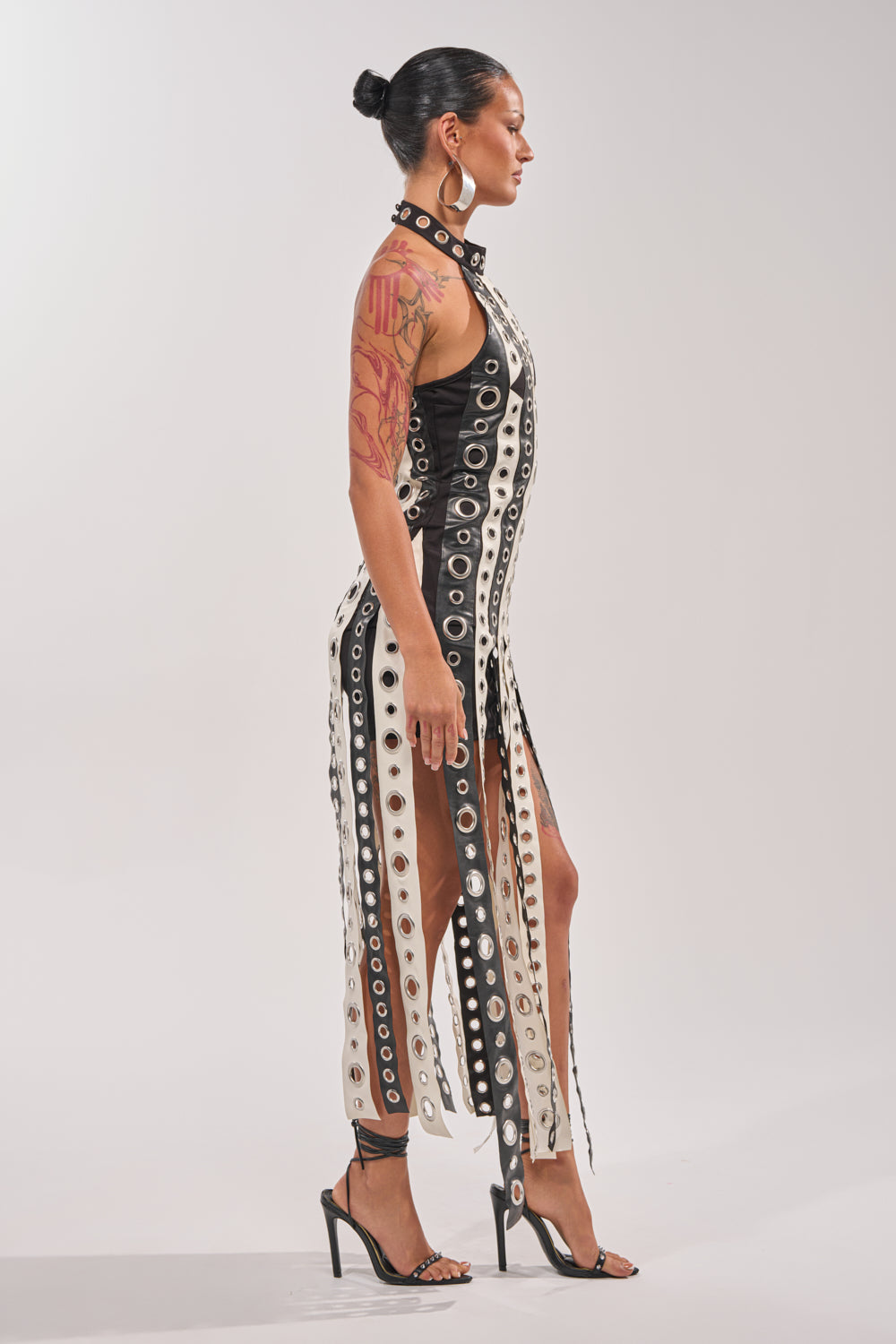 A woman in profile wears the ON MY BEST BEHAVIOR TASSEL MAXI DRESS with black high-heeled sandals. Her hair is styled in a sleek bun, and a visible arm tattoo completes the look. 3