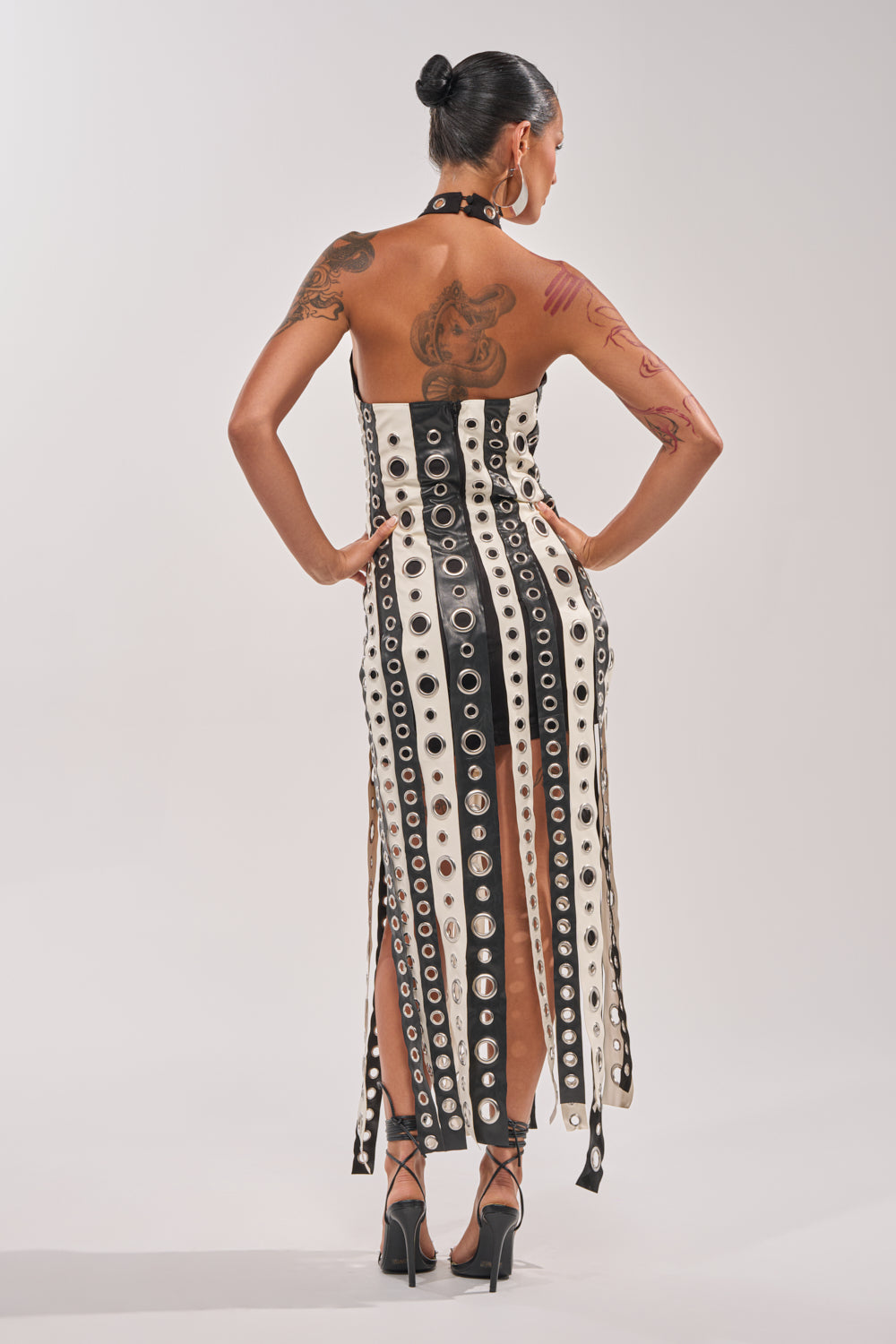 A woman with dark hair in a bun, tattoos on her arms, stands facing away with hands on hips, wearing the ON MY BEST BEHAVIOR TASSEL MAXI DRESS and black high heels. 4