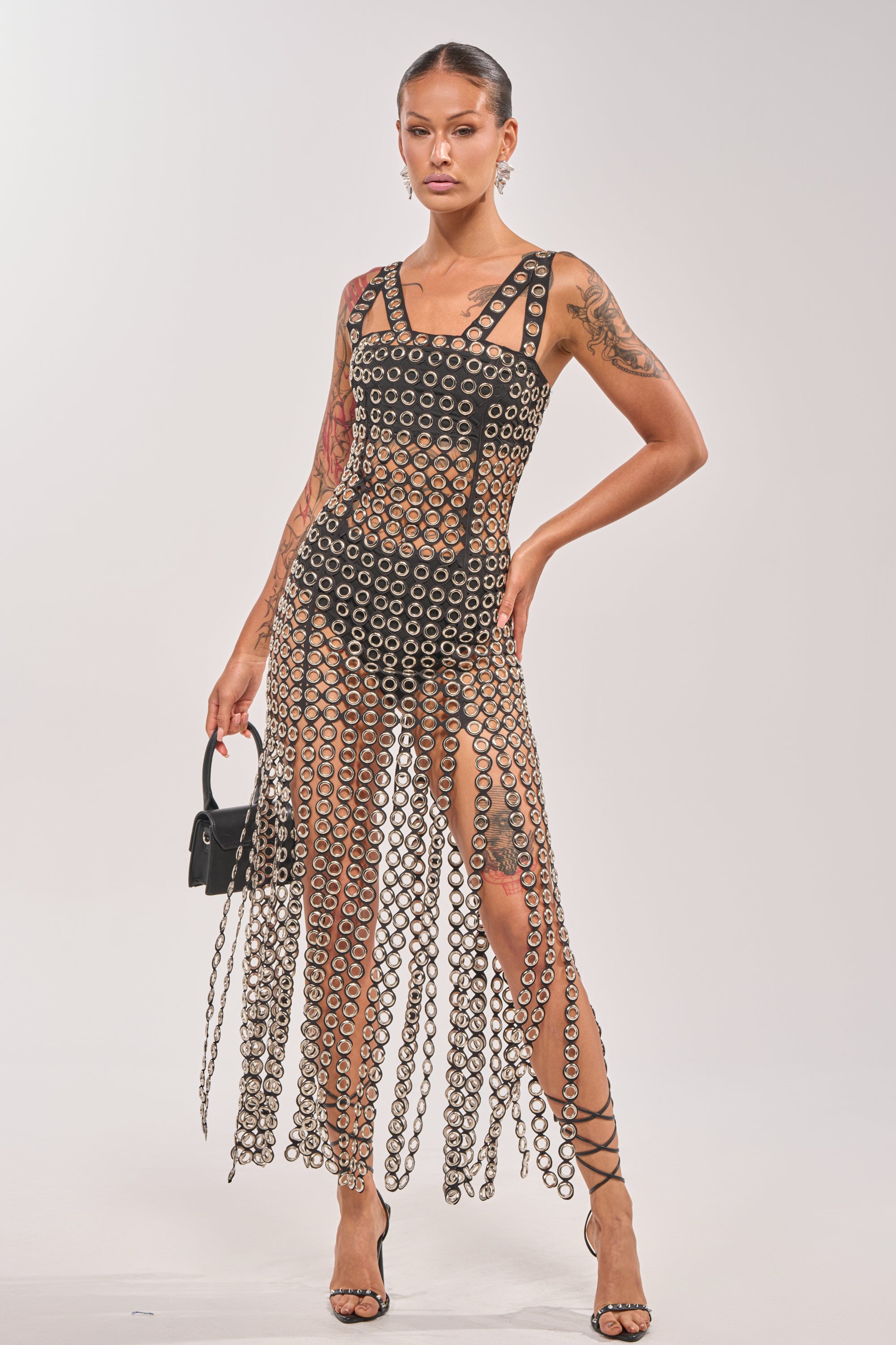 A woman models the FLIRT ALERT CHAIN MIDI DRESS, a sleeveless see-through fringe dress with metallic rings and a high slit, worn over a black bodysuit. She accessorizes with a small black handbag and strappy heels. 0