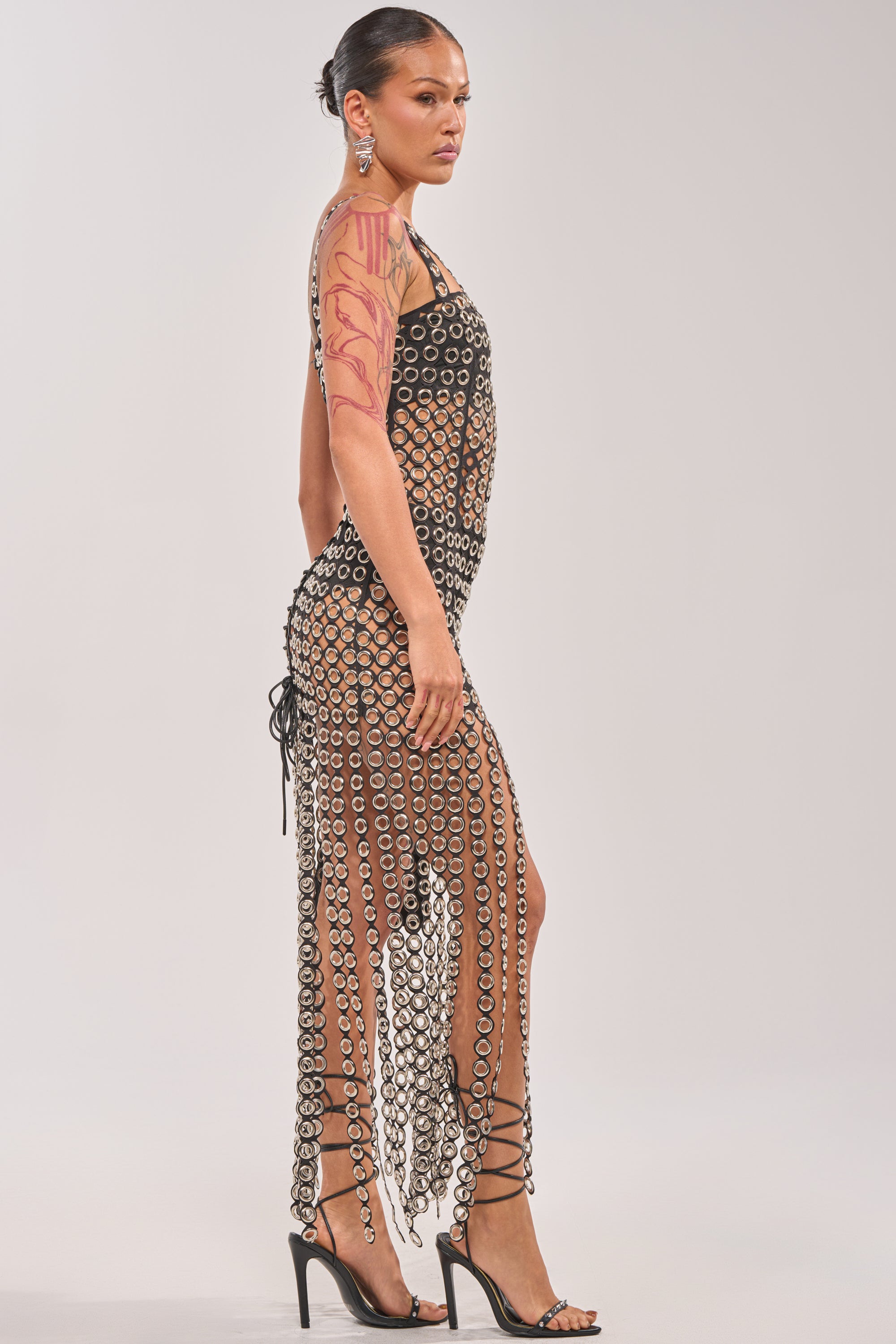 A woman stands in profile wearing the FLIRT ALERT CHAIN MIDI DRESS with circular cutouts, strappy black heels, bold earrings, a slicked-back hairstyle, and a pink abstract arm design—an eye-catching festival look. 3