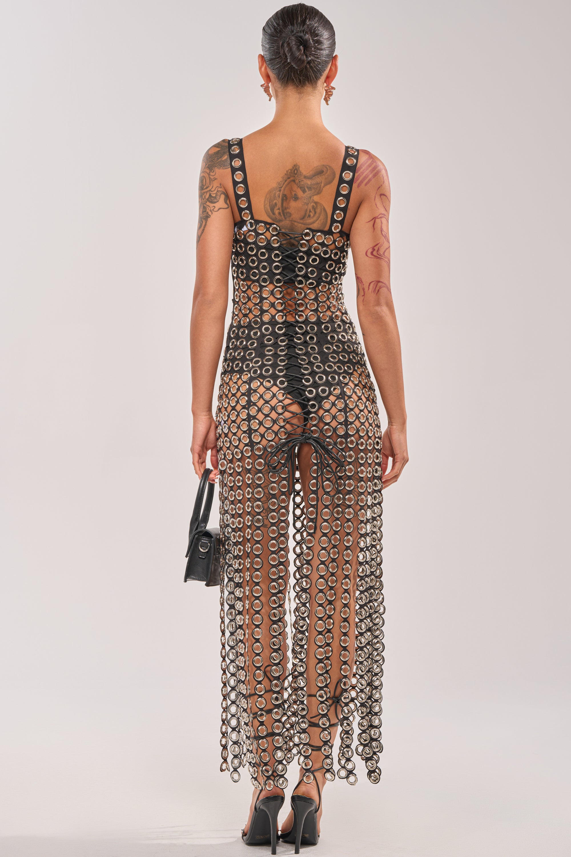 Wearing the FLIRT ALERT CHAIN MIDI DRESS with black high heels and a small black handbag, a tattooed individual stands with their back to the camera against a light background—a daring festival outfit. 4