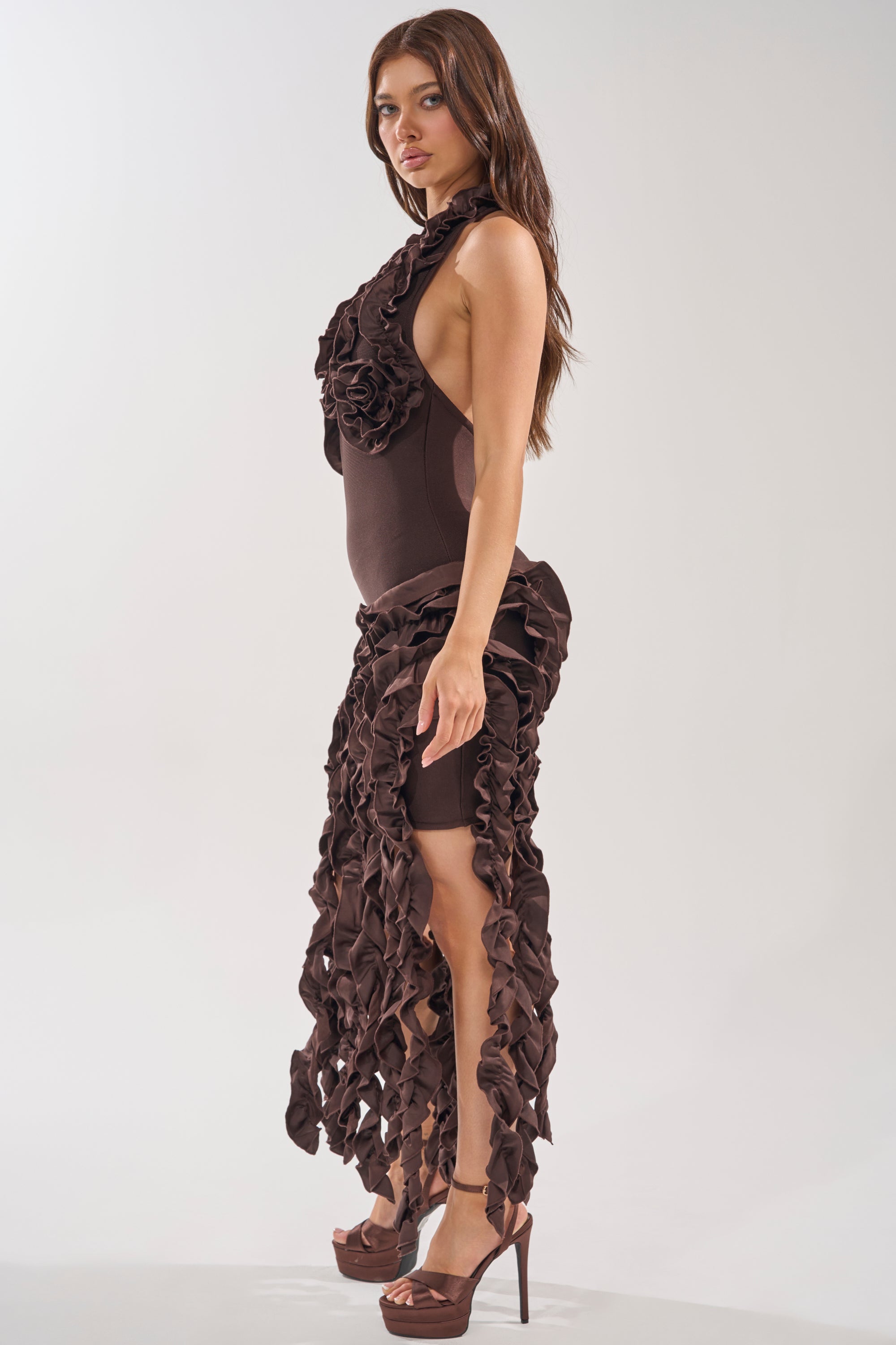 A woman models the STANDING OVATION FLOCKED MAXI DRESS IN CHOCOLATE—featuring ruffled halter style, textured details, and a high slit—paired with high platform heels against a plain light background, perfect for evening events. 2