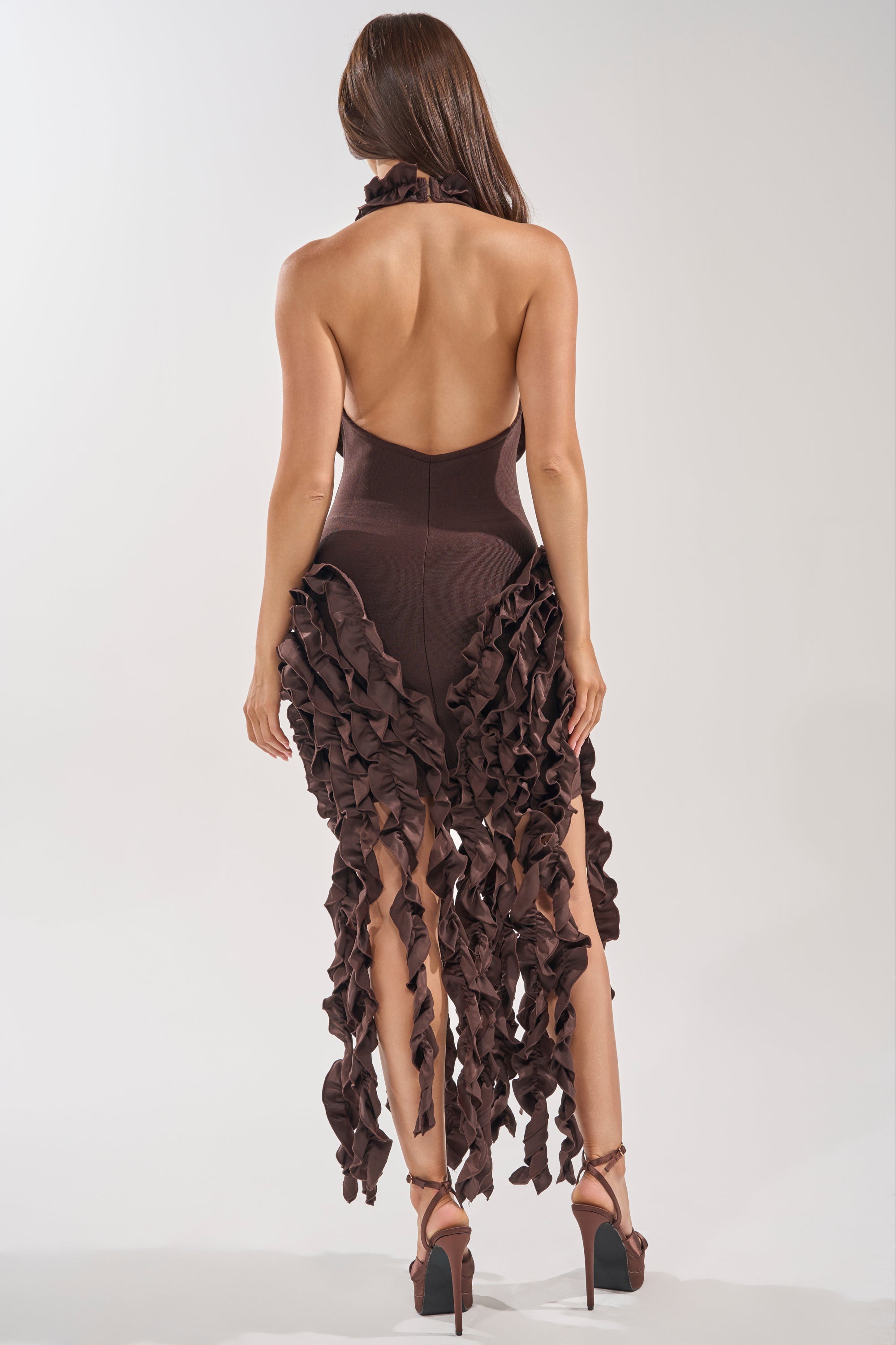 A woman faces away, wearing the STANDING OVATION FLOCKED MAXI DRESS IN CHOCOLATE with ruffled details and high-heeled sandals. The light background highlights this elegant, brown evening maxi dress. 3