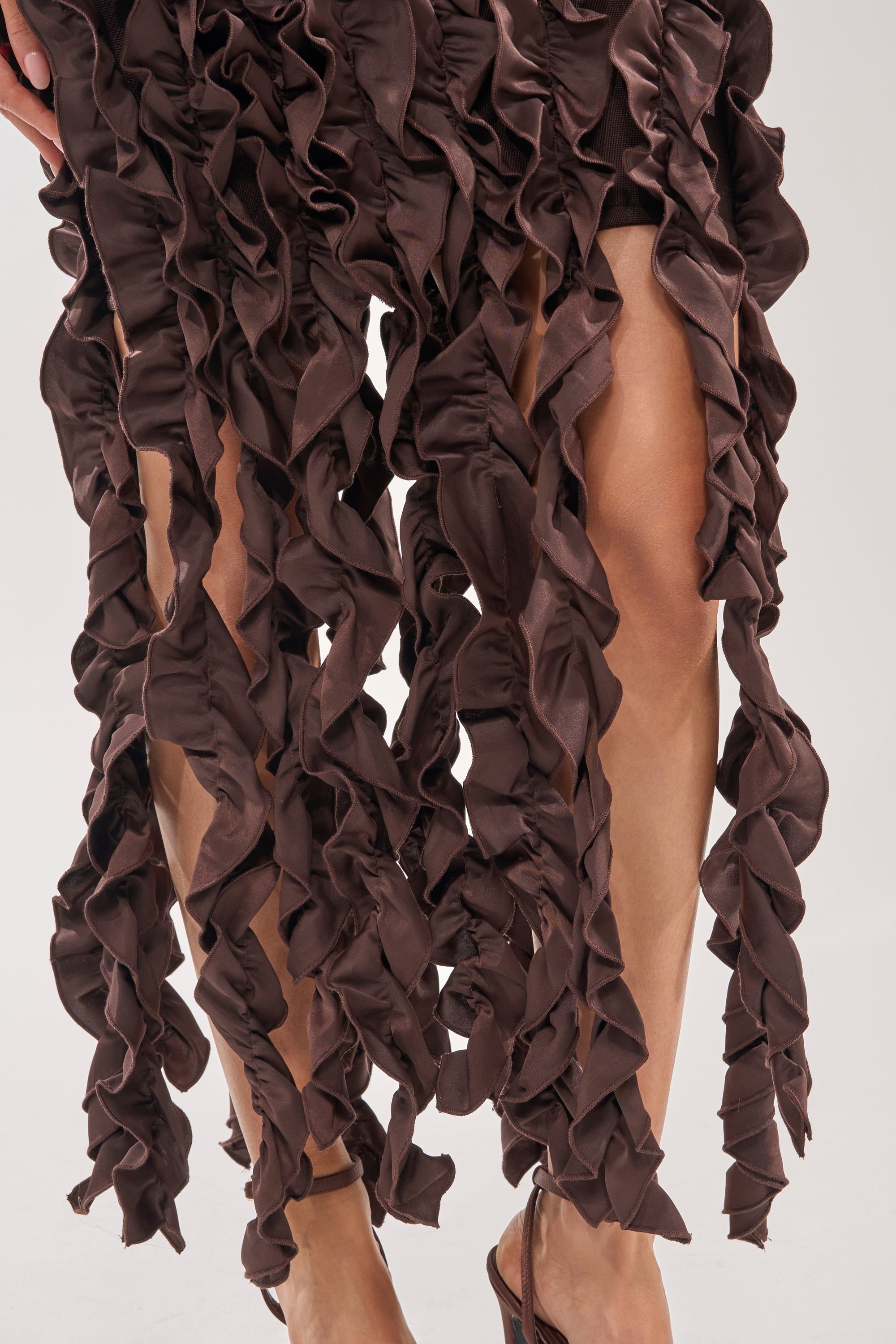 Close-up of a person in the STANDING OVATION FLOCKED MAXI DRESS IN CHOCOLATE, featuring long ruffled detailing and a maxi silhouette that reveals their legs and strappy sandals—ideal for an elegant evening occasion. 6