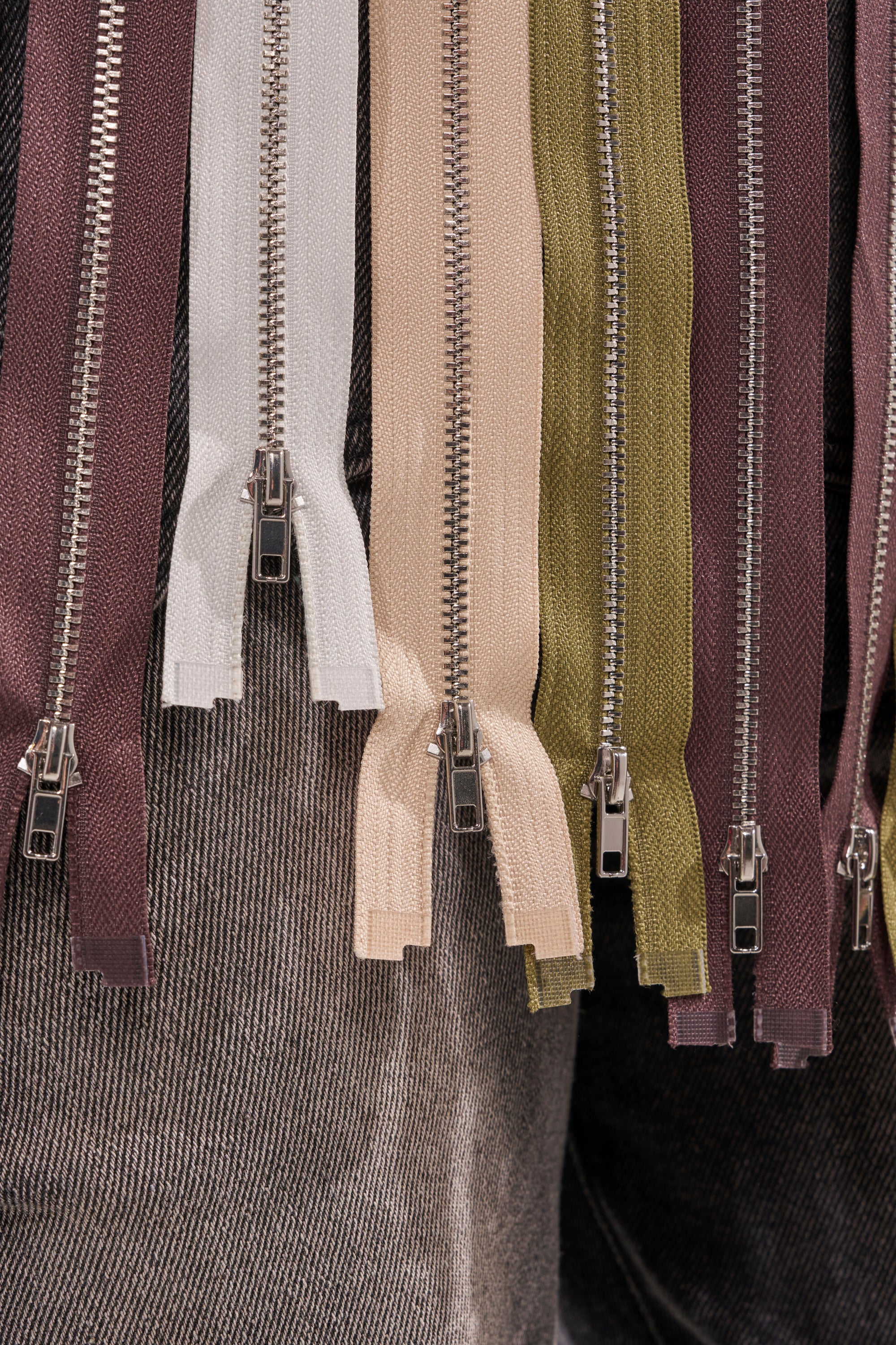 Close-up of multiple colorful zippers—brown, white, beige, and olive—hanging over gray denim fabric with metal pulls visible, reflecting the bold style of the ZIPPED THROUGH SLEEVELESS TOP. 11