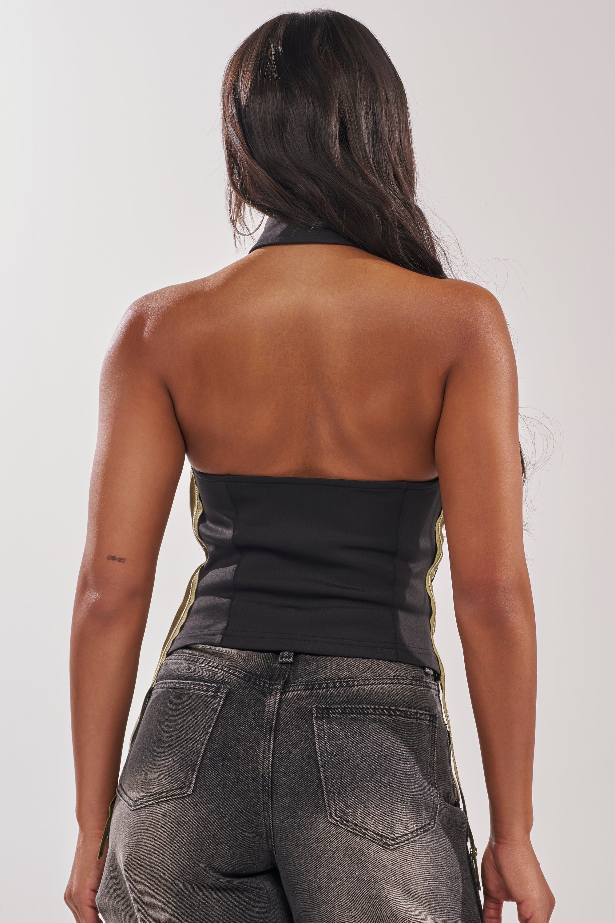 A woman with long dark hair faces away, wearing the ZIPPED THROUGH SLEEVELESS TOP and faded gray high-waisted jeans. Her back and shoulders are visible against a neutral background—ideal for chic nightlife style. 7