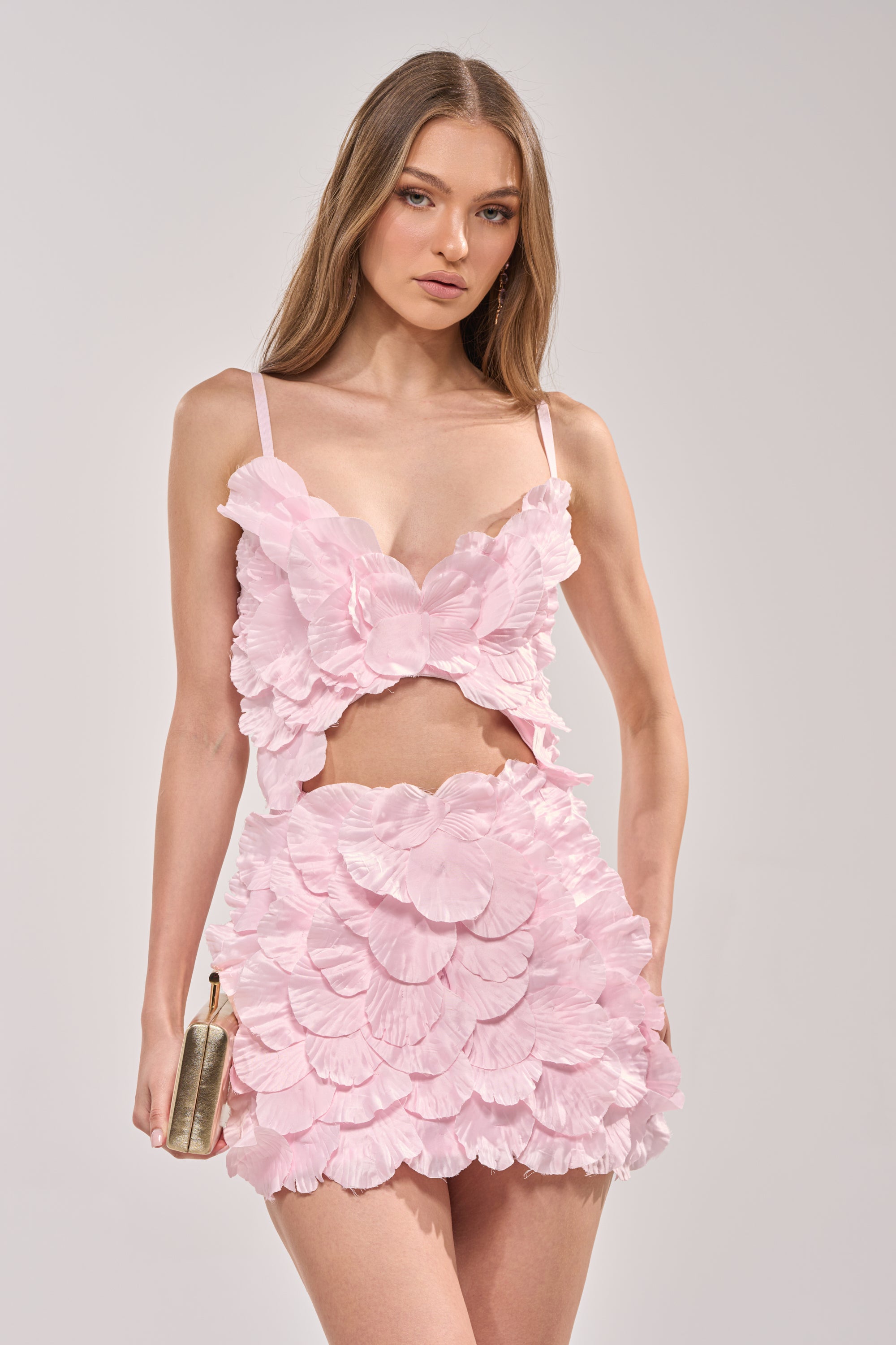 A woman wears the CAUGHT YOUR ATTENTION FLORAL APPLIQUE MINI DRESS, a light pink, petal-textured mini dress. She holds a gold clutch, has long straight hair, and stands against a simple light background. 3
