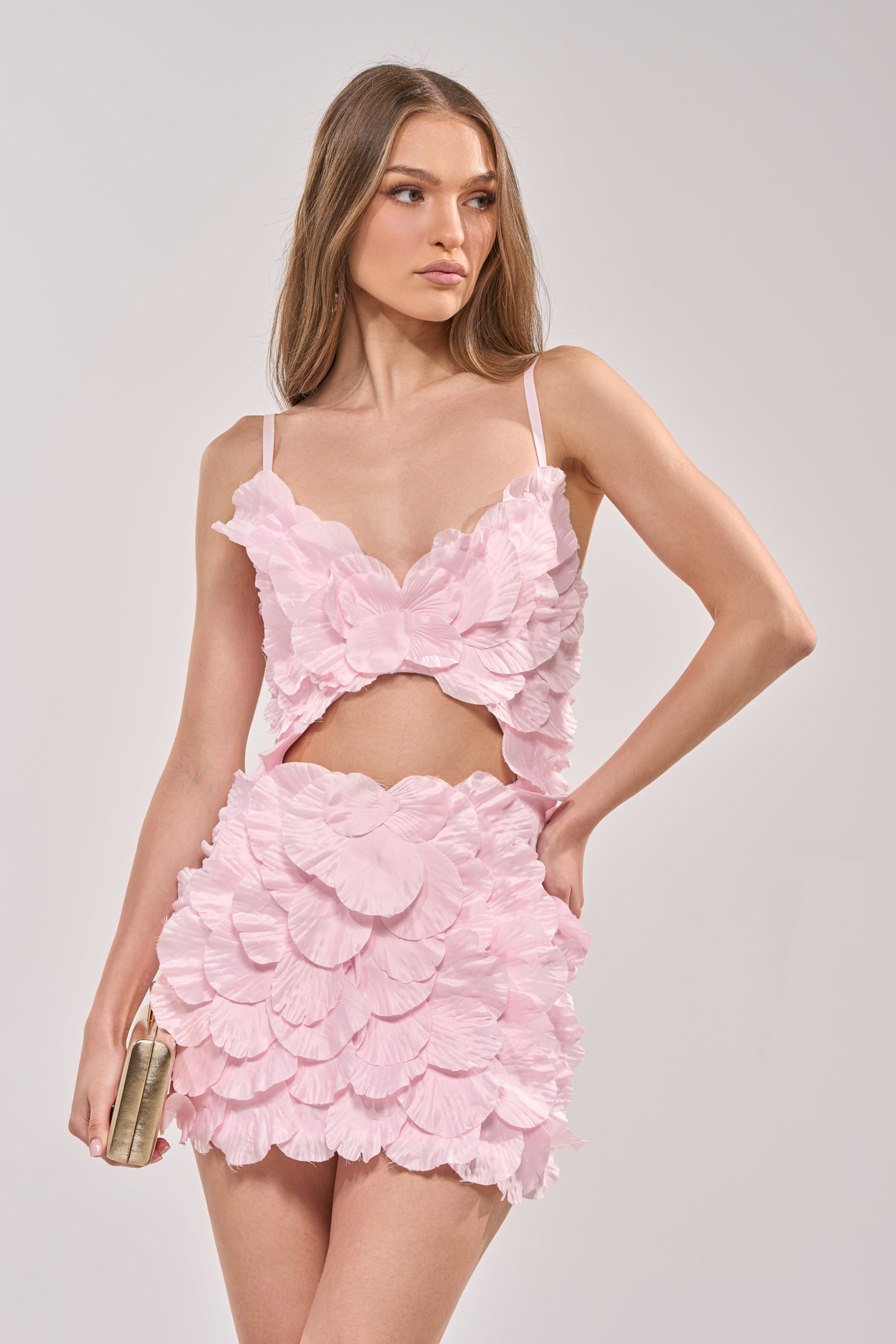 A woman in the CAUGHT YOUR ATTENTION FLORAL APPLIQUE MINI DRESS—a blush pink, sleeveless bodycon mini with layered floral ruffles—holds a small gold clutch against a plain light background. 4