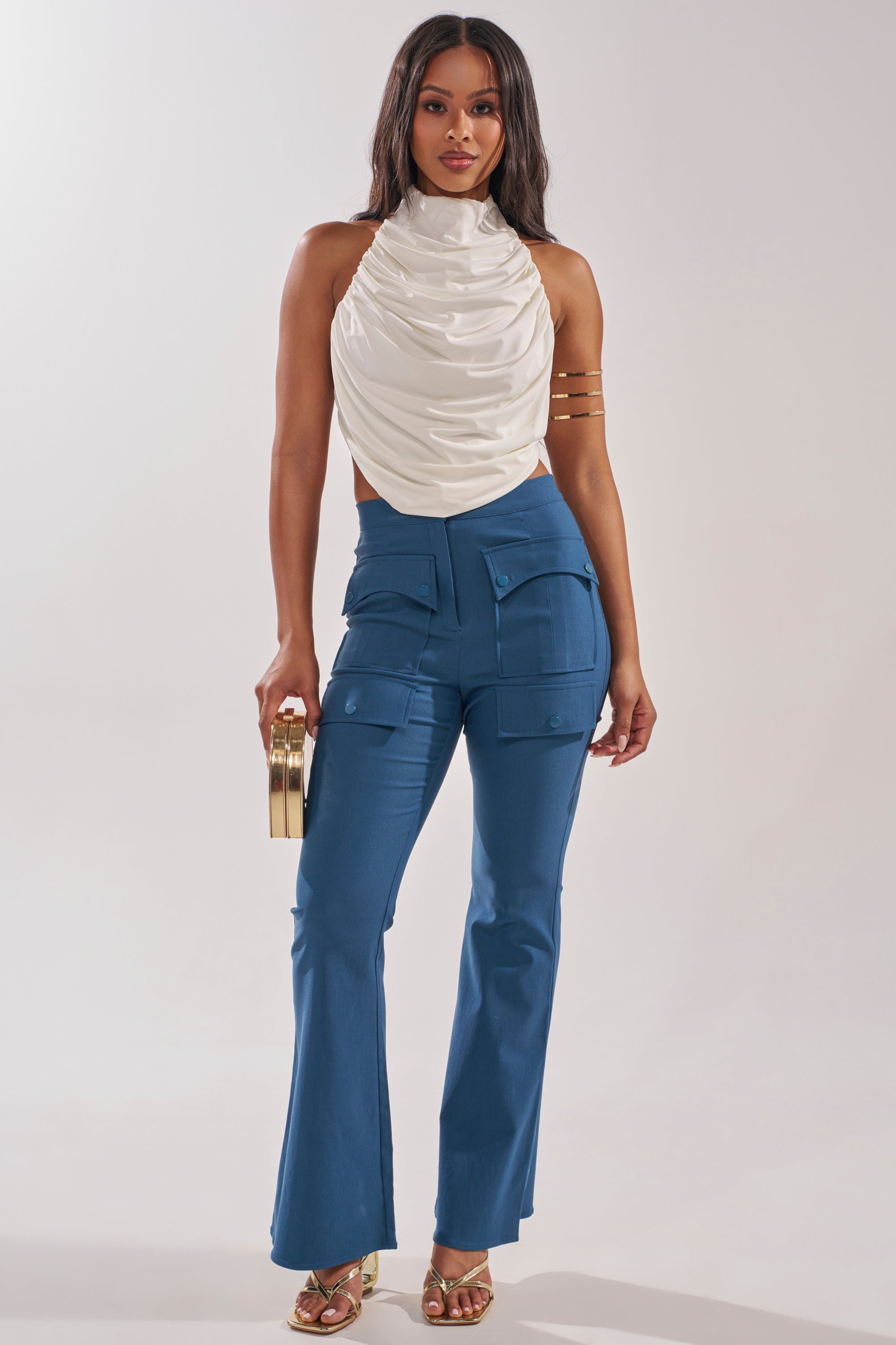 A woman poses against a plain background wearing a sleeveless white draped top, the TRY ME TROUSER blue high-rise flared utility pants with cargo pockets, gold heels, an arm cuff, and a small gold handbag. 0
