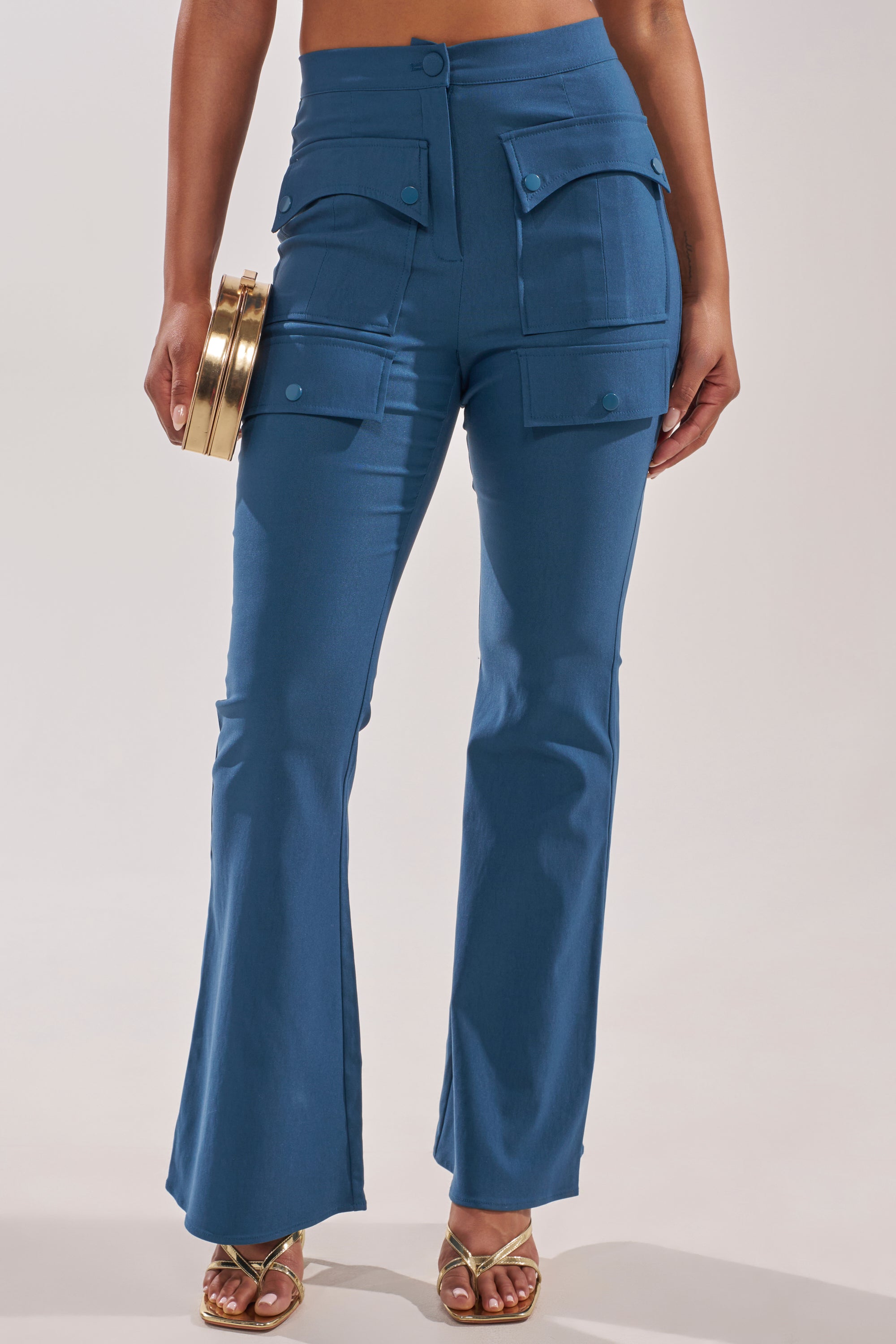 Wearing the TRY ME TROUSER—high-rise blue flared utility pants with cargo flap pockets—paired with gold sandals and a round gold clutch; image spotlights the lower torso and legs. 1