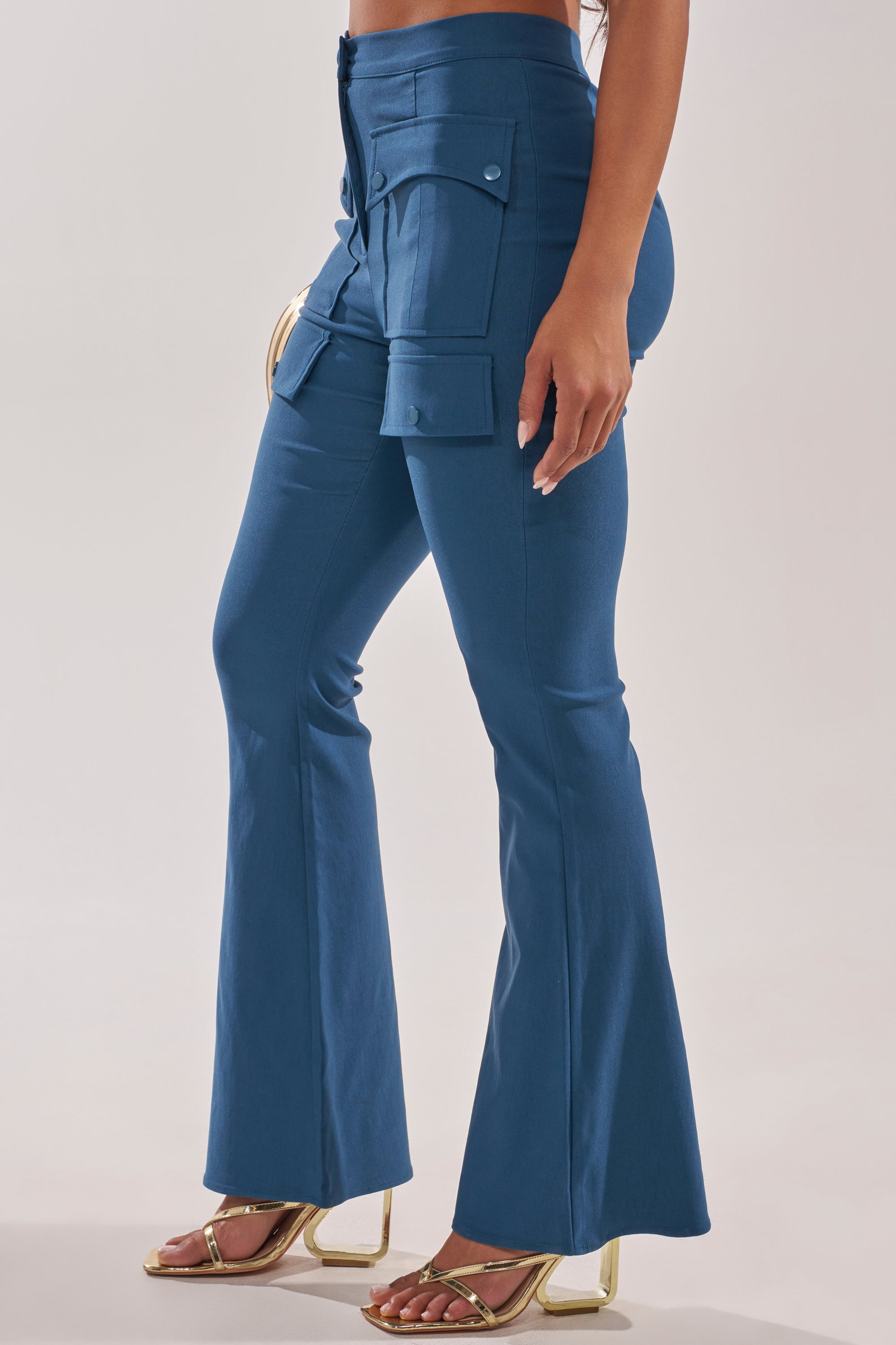 A woman stands against a plain background wearing the TRY ME TROUSER—blue, high-rise flared utility trousers with large cargo flap pockets—paired with gold high-heeled sandals. 3