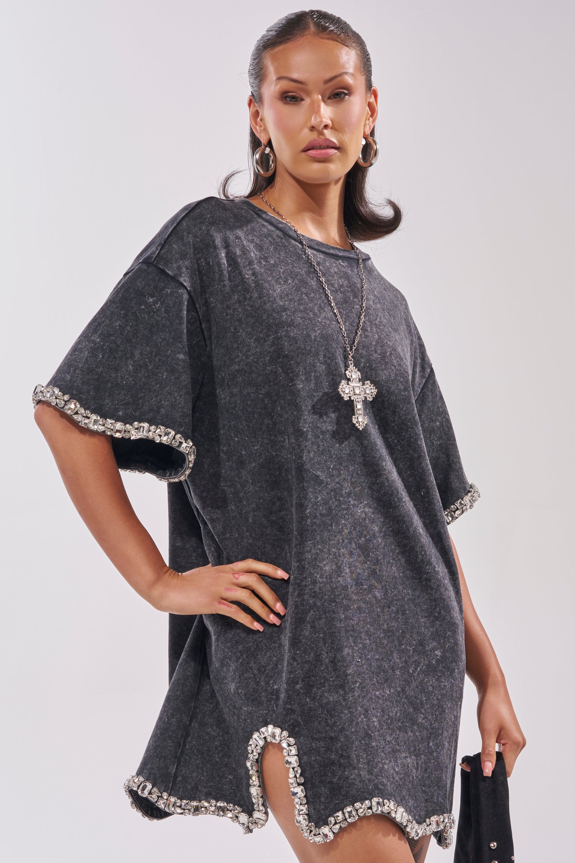 A woman poses confidently in the COOL GIRL EMBELLISHED TEE SHIRT DRESS IN BLACK, a loose washed black mini dress with rhinestone embellishments on the sleeves and hem, styled with hoop earrings and a bold cross necklace against a light background. 1
