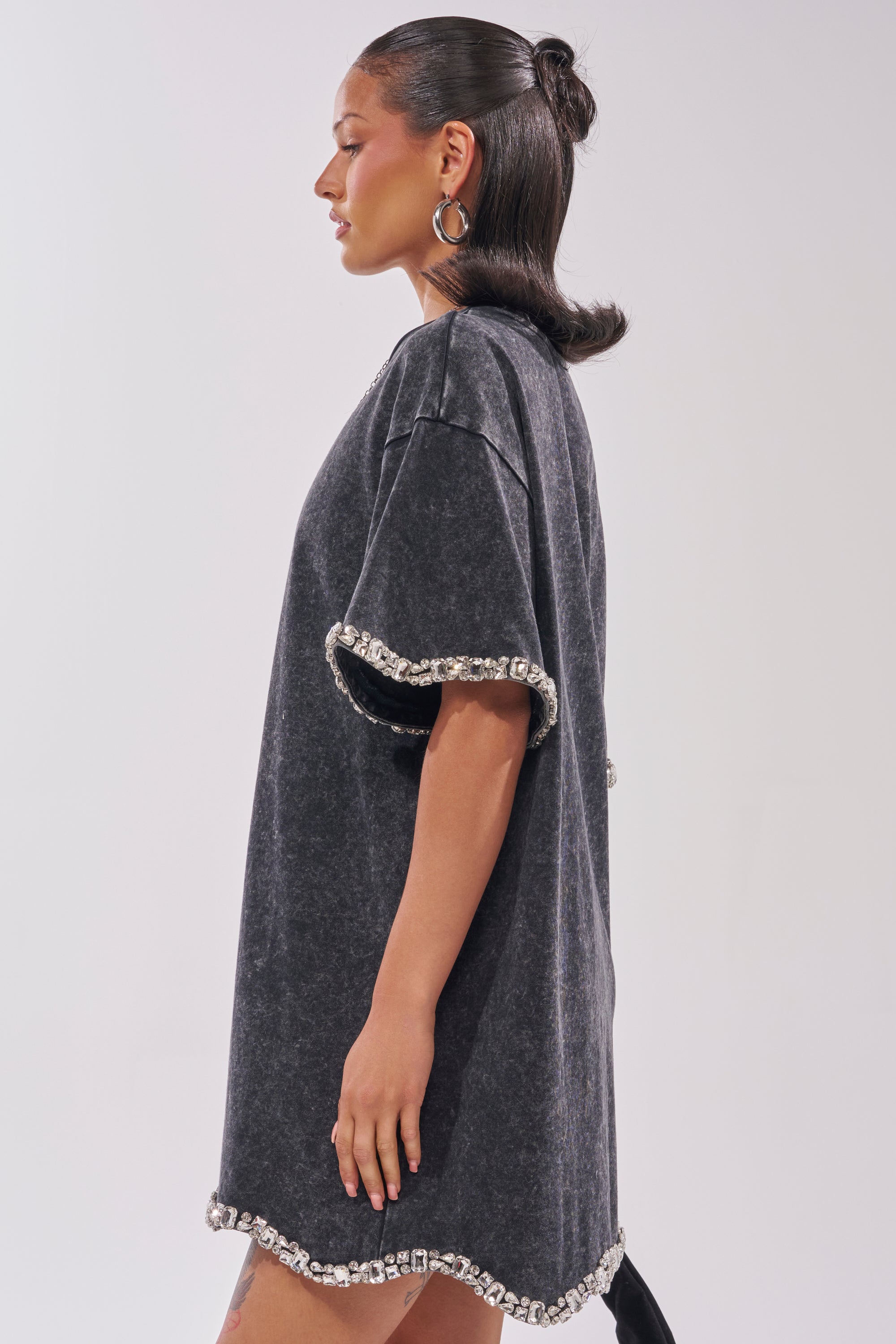 A woman stands in profile wearing the COOL GIRL EMBELLISHED TEE SHIRT DRESS IN BLACK, featuring large silver rhinestone trim on the sleeves and hem—her sleek hair and hoop earrings complete this chic date night look against a neutral background. 4