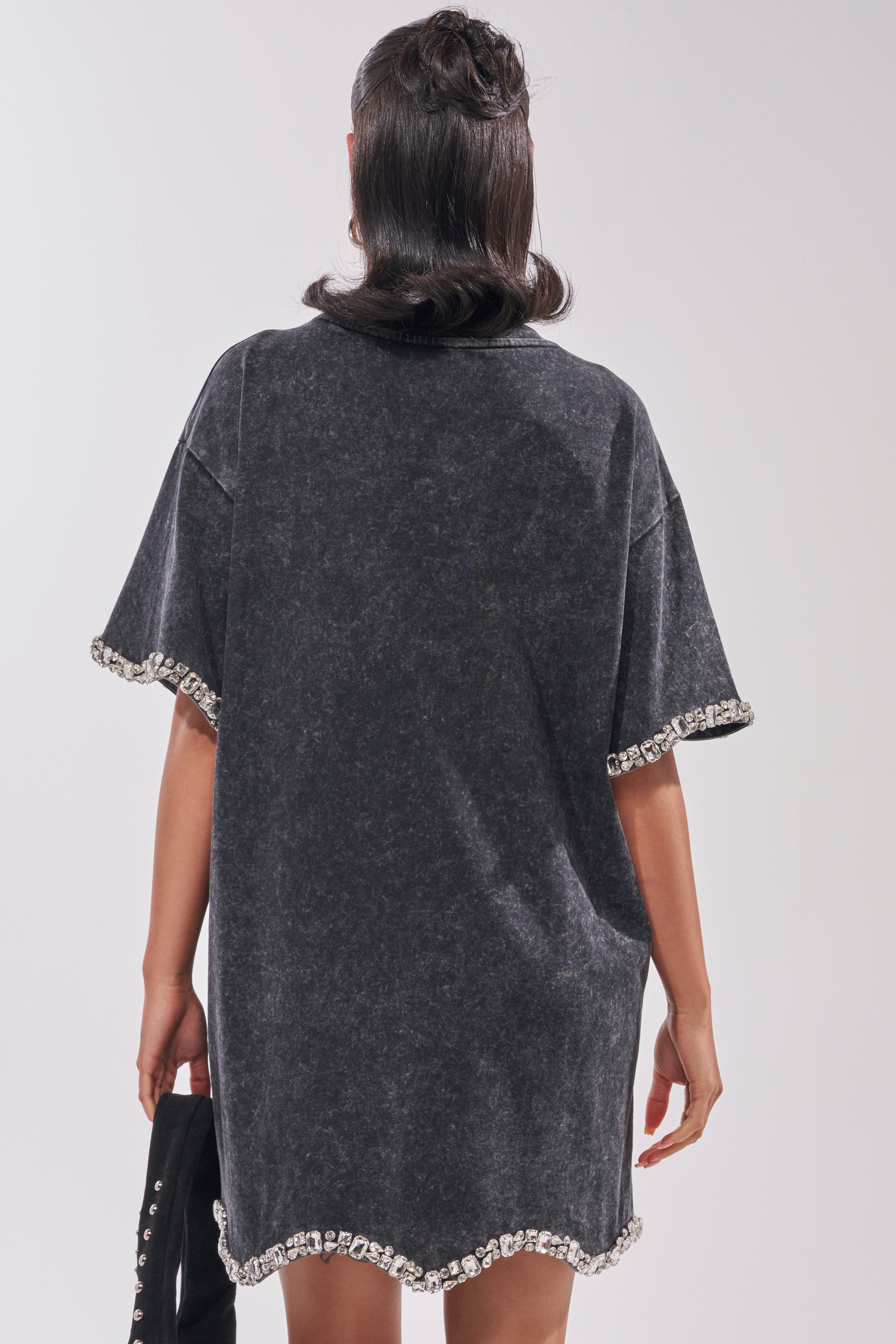 A person with shoulder-length dark hair is seen from the back wearing the COOL GIRL EMBELLISHED TEE SHIRT DRESS IN BLACK, a loose-fit, short-sleeve mini dress adorned with decorative rhinestones along the sleeves and hem. 5