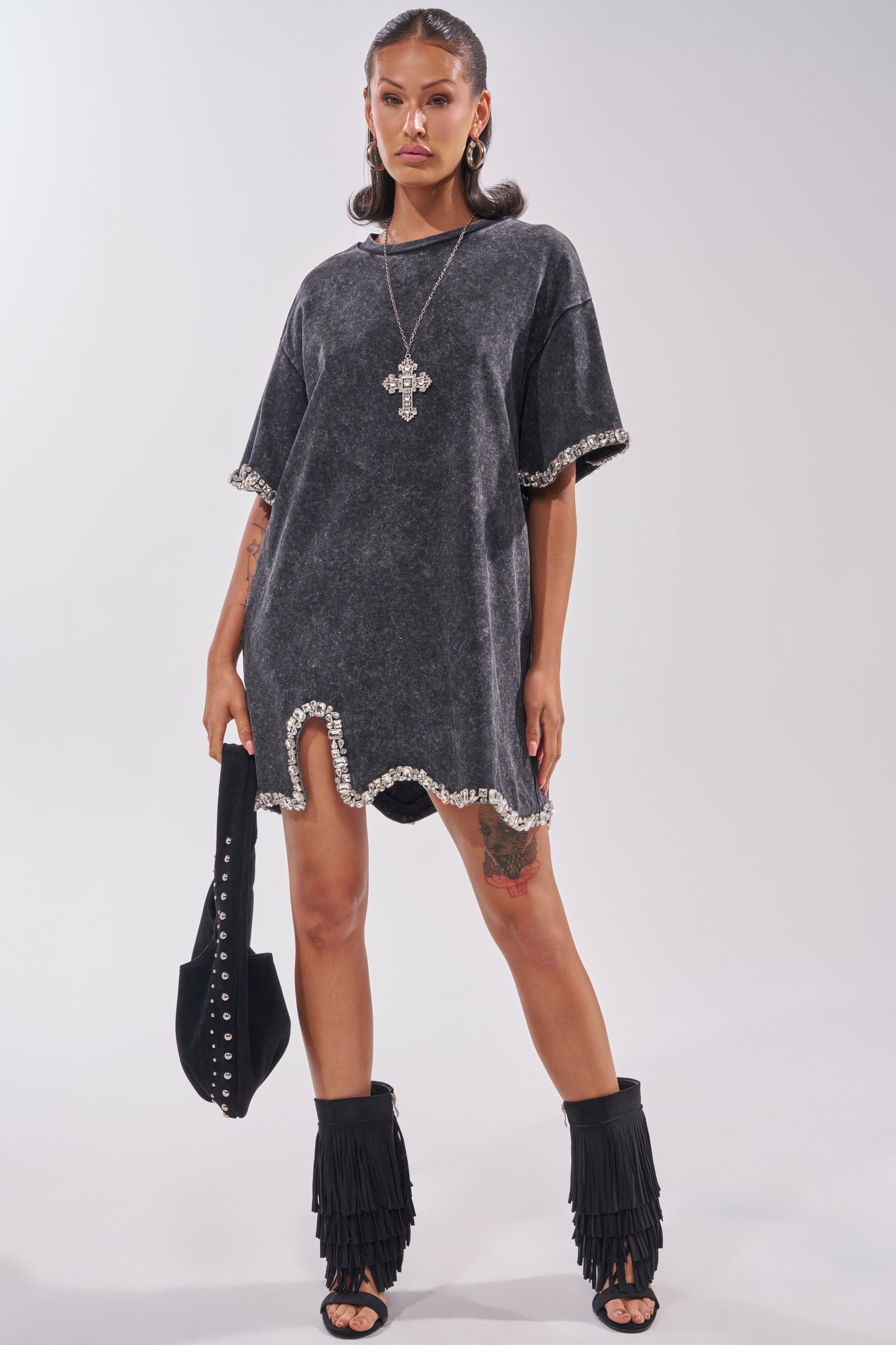 A woman stands confidently in the COOL GIRL EMBELLISHED TEE SHIRT DRESS IN BLACK, paired with black fringe ankle boots, a bold cross necklace, a studded bag, slicked-back hair, and visible tattoos for the perfect date night statement look. 6