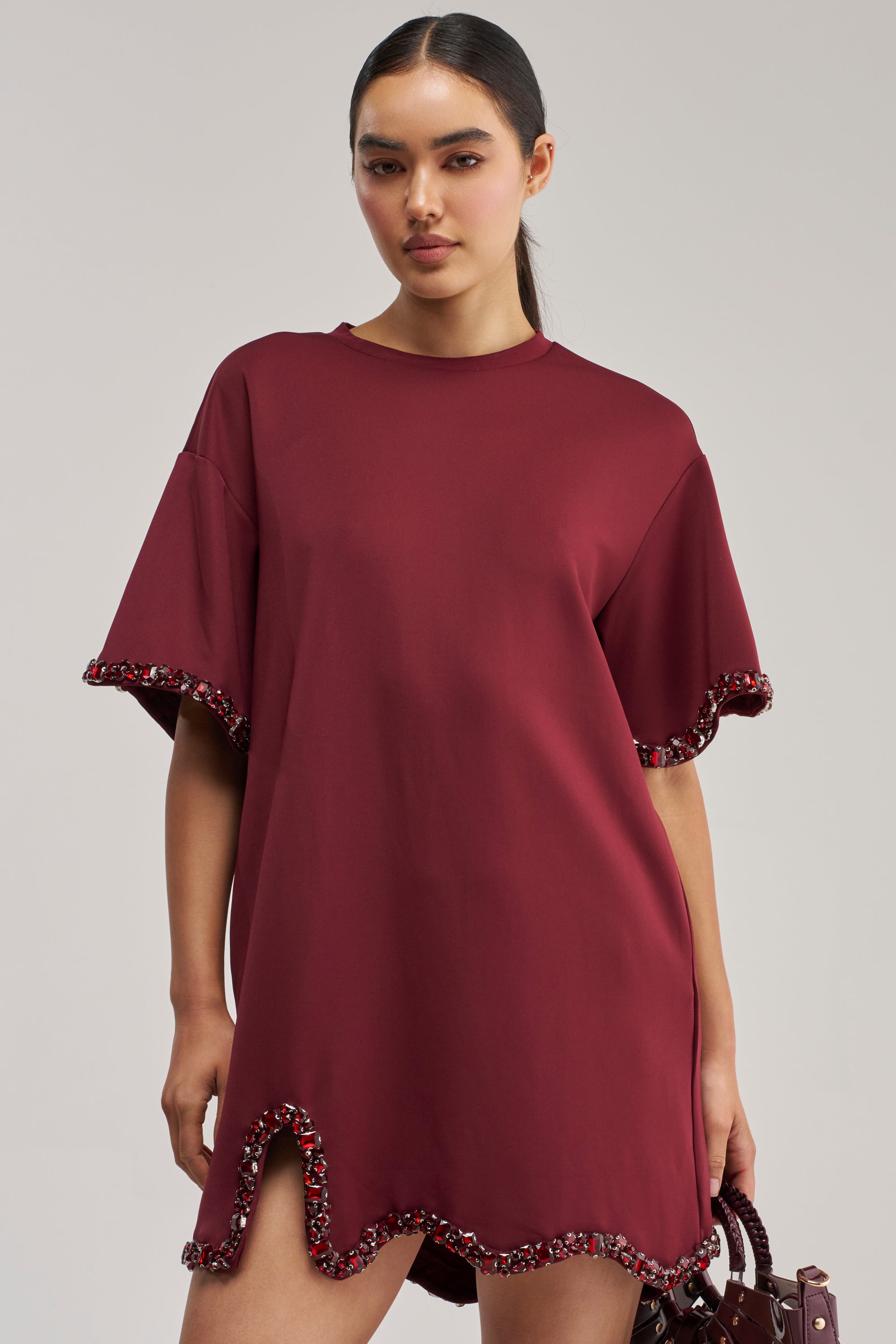 A woman models the COOL GIRL EMBELLISHED TEE SHIRT DRESS IN BURGUNDY, a rhinestone-adorned dress featuring short sleeves, a high-low hem, and a side slit. The sleeve and hem edges are embellished with dark red faceted crystals for extra sparkle, making it perfect for a glamorous night out. She completes the look with a matching burgundy handbag against a light gray background.