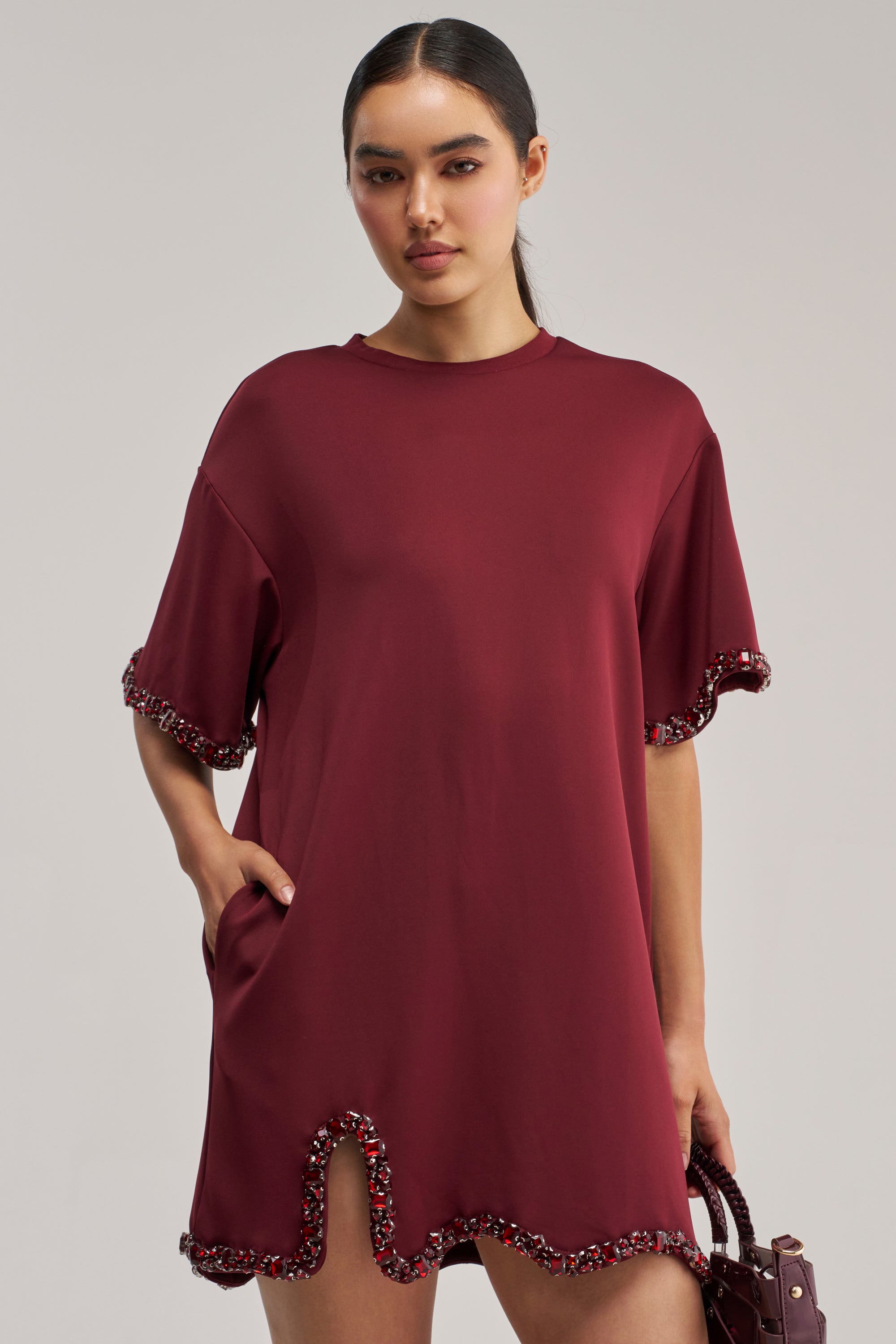 A woman poses against a plain background wearing the COOL GIRL Embellished Tee Shirt Dress in Burgundy, a short-sleeved mini dress featuring beaded trim at the sleeves and hem. She stands with one hand in her pocket and holds a small bag in the other.