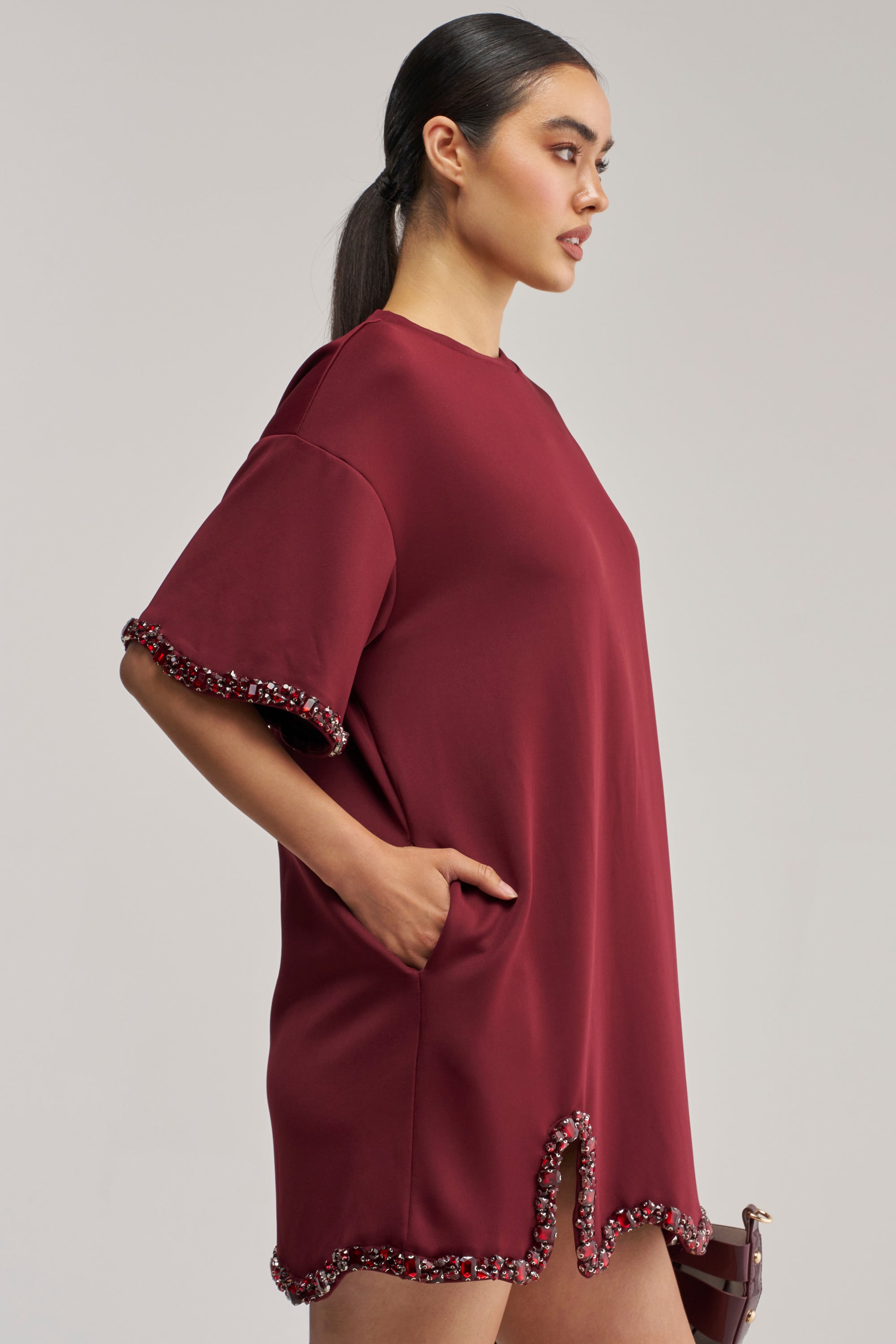 A woman wearing the COOL GIRL Embellished Tee Shirt Dress in Burgundy, standing in profile with one hand in her pocket, hair tied back, holding a bag, against a plain light background.