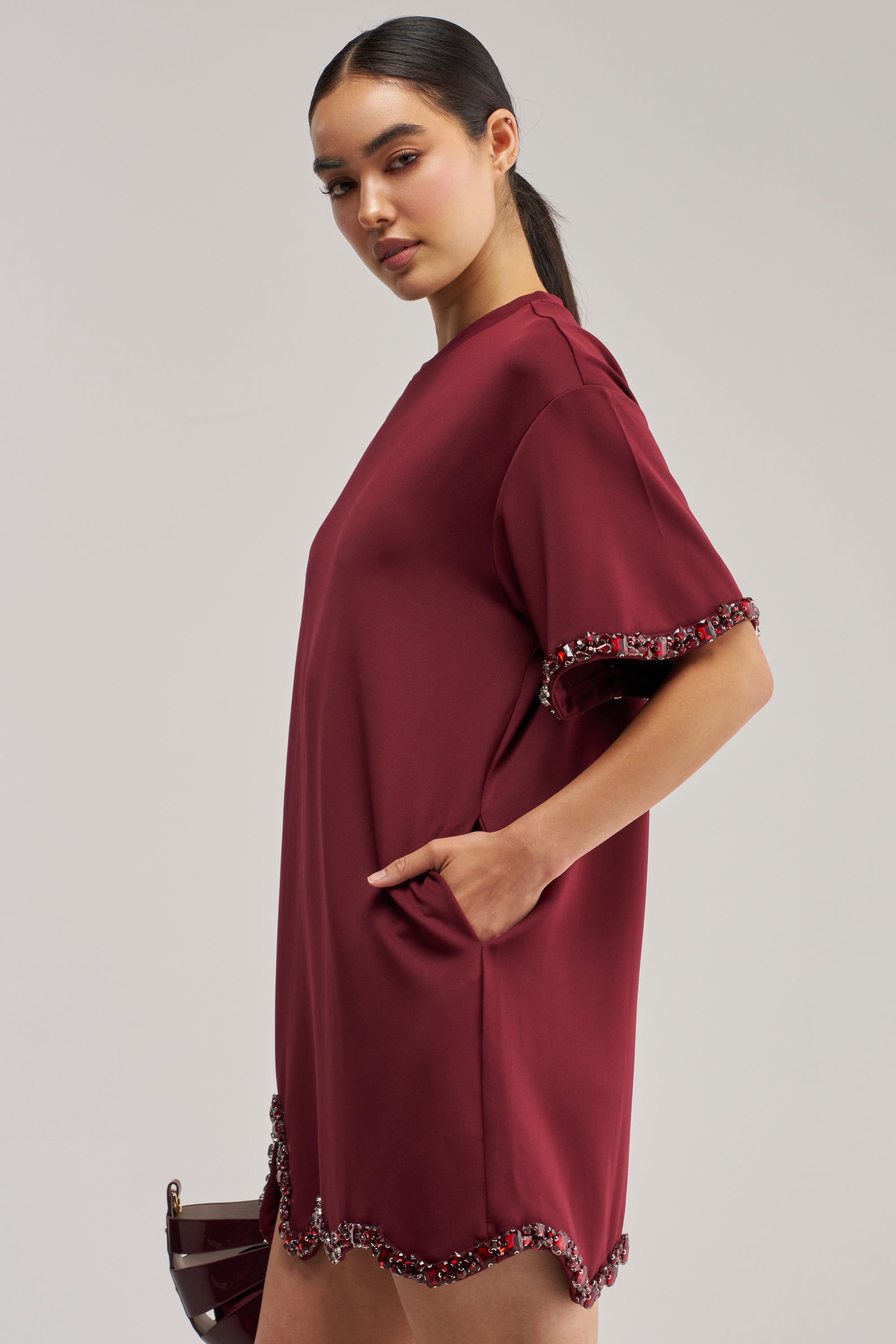 A model showcases the COOL GIRL EMBELLISHED TEE SHIRT DRESS IN BURGUNDY, a mini dress adorned with jeweled crystal embellishments along the hem and sleeves. She poses with one hand in the pocket and holds a matching clutch—an ideal choice for a birthday dress or to elevate your style with the sparkle of a rhinestone T-shirt dress.
