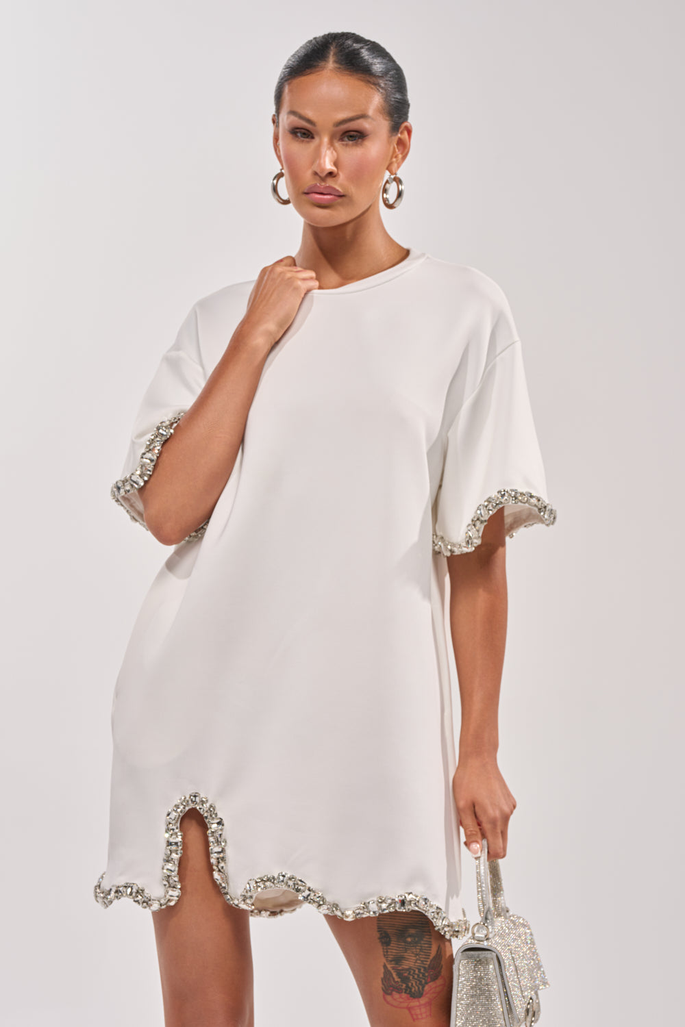 A woman in the COOL GIRL EMBELLISHED TEE SHIRT DRESS IN WHITE with jeweled trim and sleeves holds a sparkly handbag. She has hoop earrings, a sleek hairstyle, and a visible tattoo on her left thigh. 0