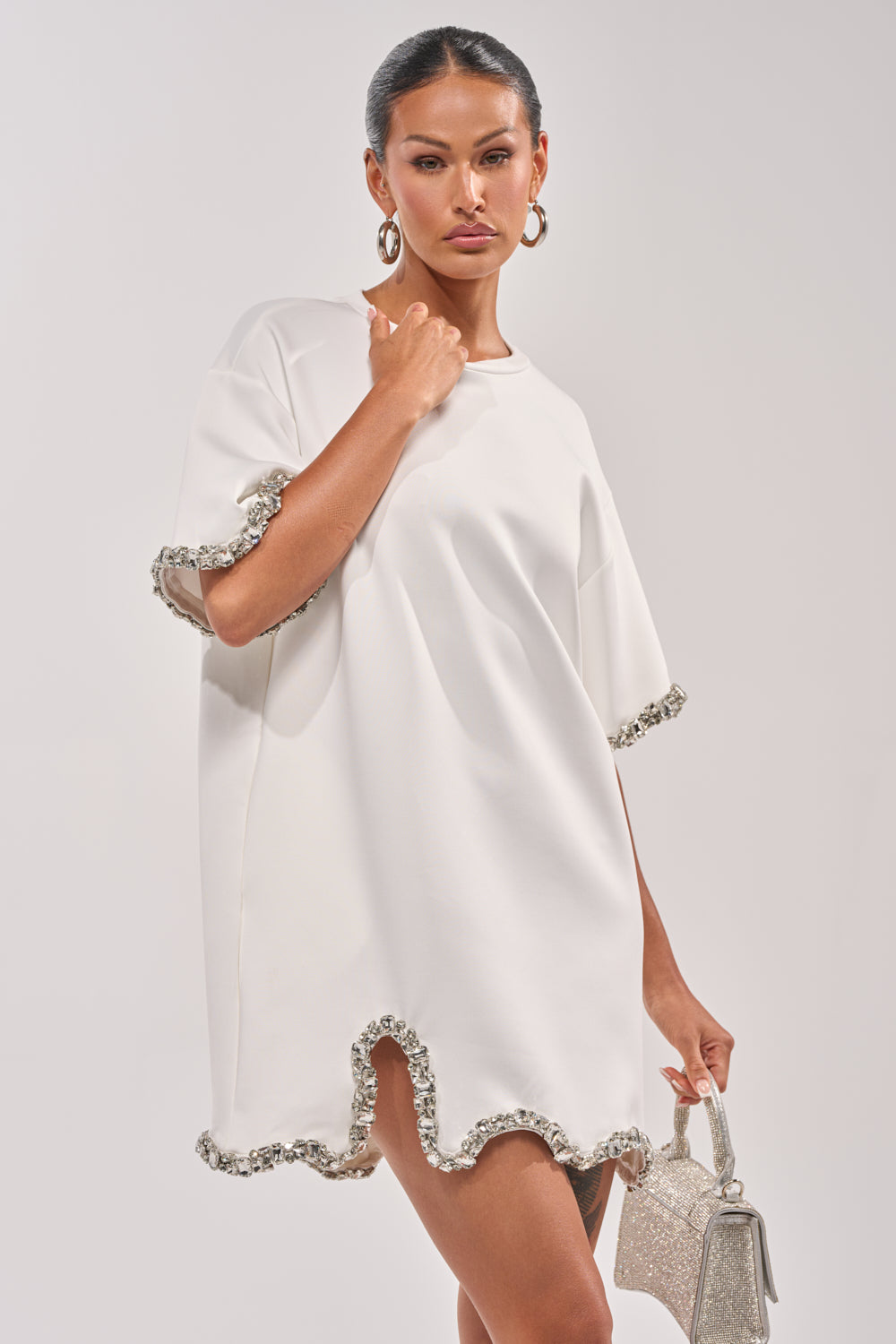 A woman wears the COOL GIRL EMBELLISHED TEE SHIRT DRESS IN WHITE with rhinestone-trimmed sleeves and hem, styled with hoop earrings and a sparkly silver handbag. Her dark hair is pulled back against a plain light background. 1