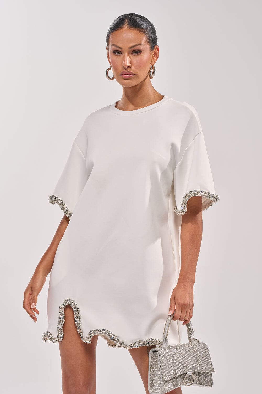 A woman models the COOL GIRL EMBELLISHED TEE SHIRT DRESS IN WHITE—a loose mini dress with ruffled edges and rhinestone trim—paired with a small silver handbag and large hoop earrings. 2