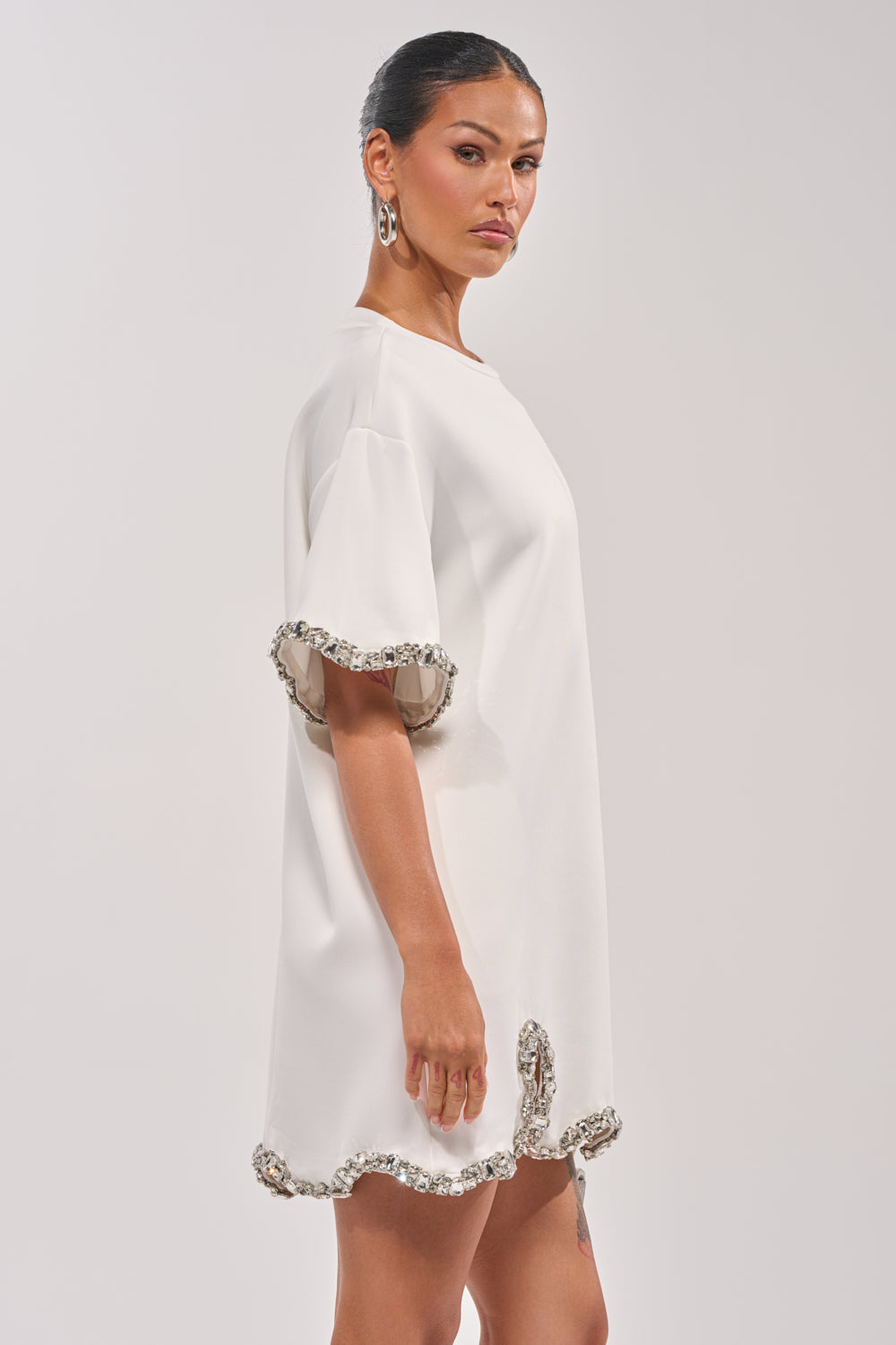 A woman stands in profile against a plain light background, dark hair pulled back and wearing hoop earrings. She models the COOL GIRL EMBELLISHED TEE SHIRT DRESS IN WHITE, a loose mini dress with short sleeves and sparkling rhinestone trim. 3